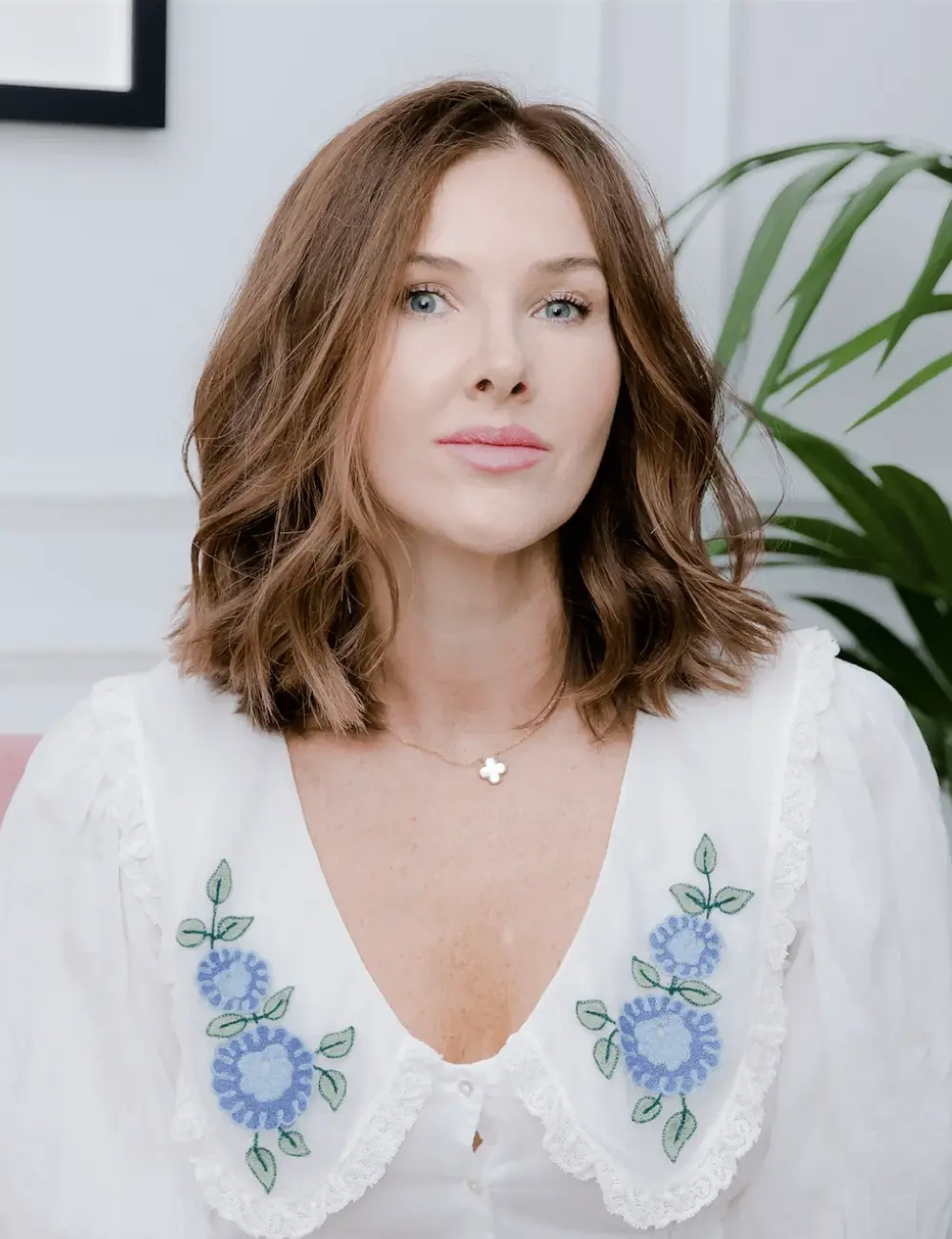 Woman with light brown shoulder-length hair wearing a white blouse with blue floral embroidery and a small flower pendant necklace.