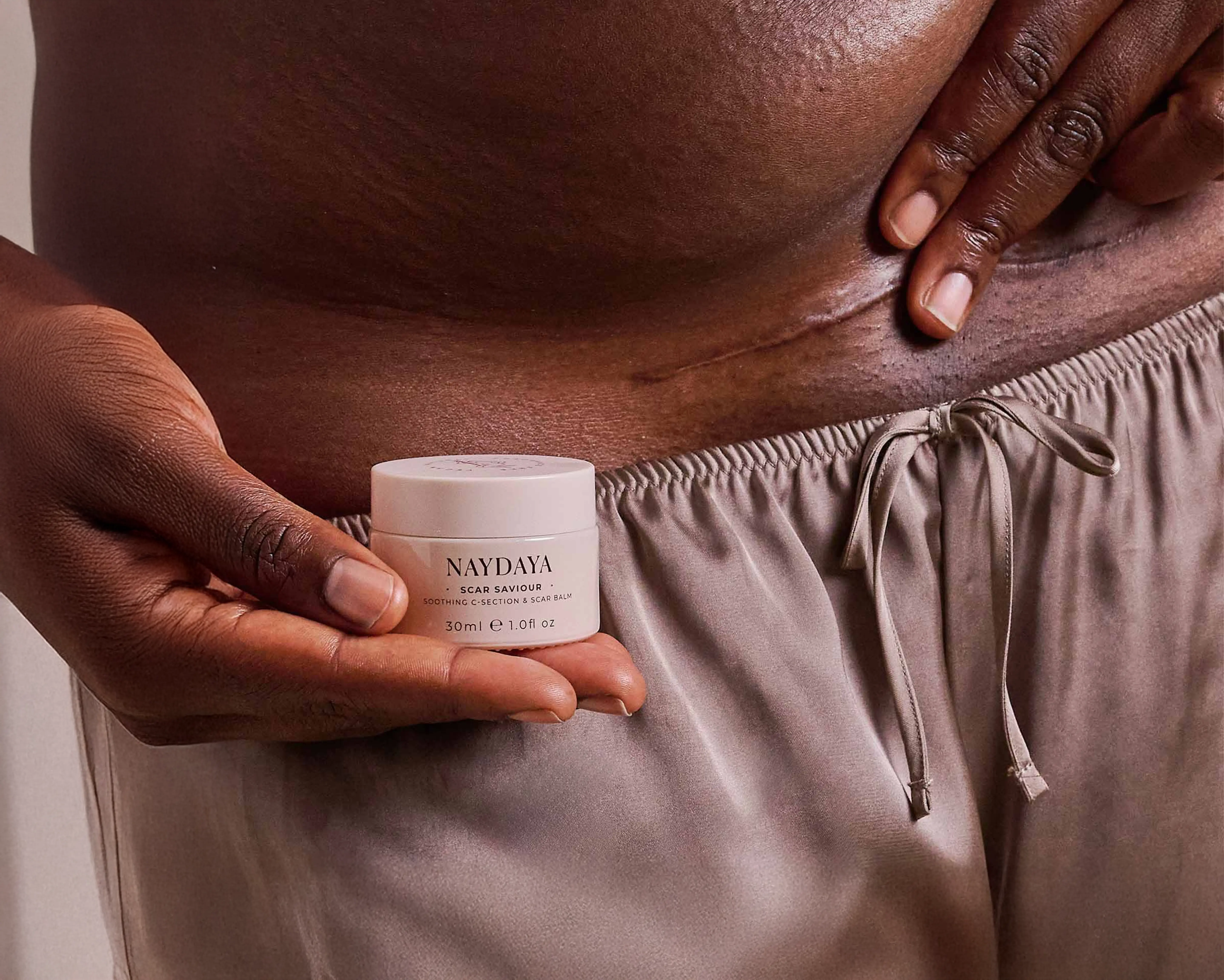 Close-up of a person holding a jar of Naydaya Scar Saviour cream near a visible C-section scar on their abdomen.