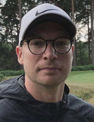 Man wearing round glasses, a white Nike cap, and a dark jacket outdoors with trees and grass in the background.
