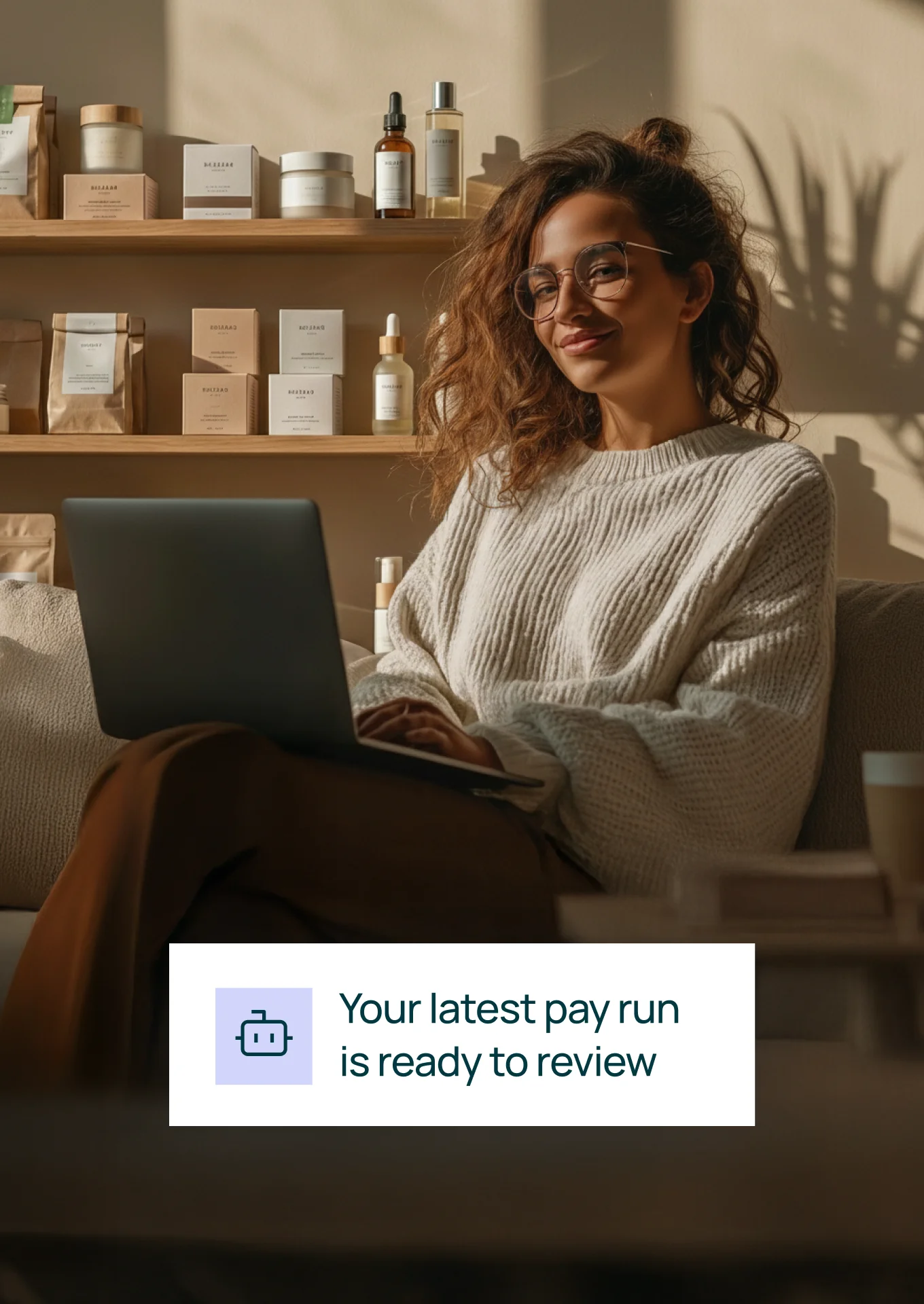 Woman wearing glasses and a cream sweater working on a laptop with shelves of skincare products in the background and text overlay reading 'Your latest pay run is ready to review'.