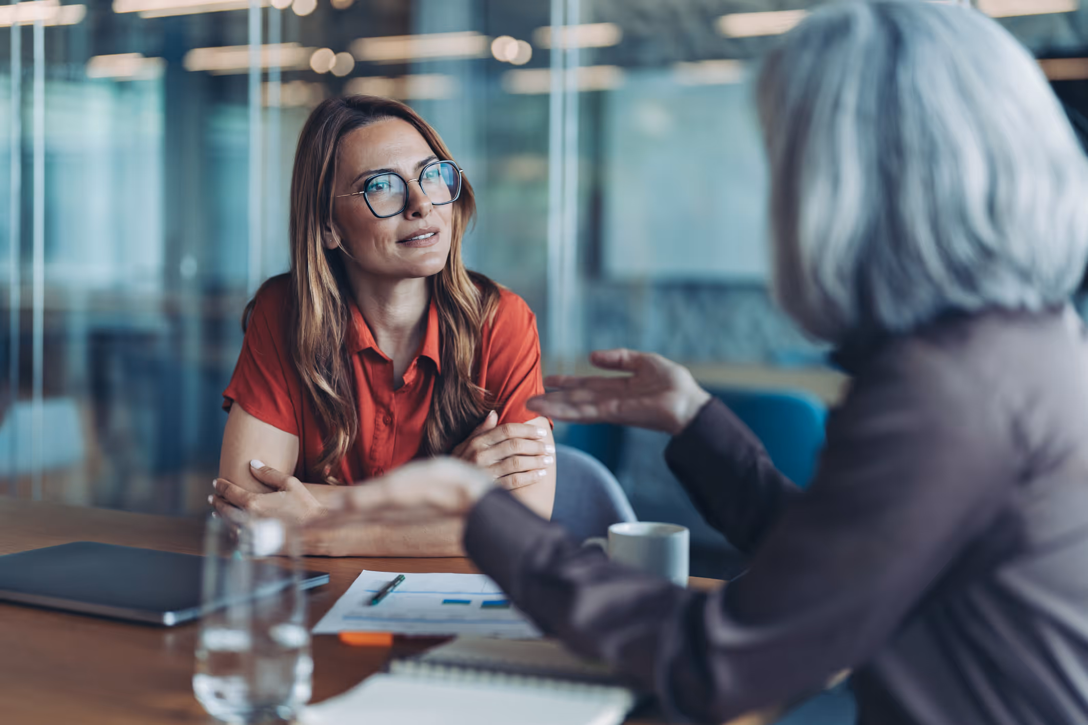 Two women having a discussion in a modern office, with one woman listening attentively wearing glasses and a red shirt.