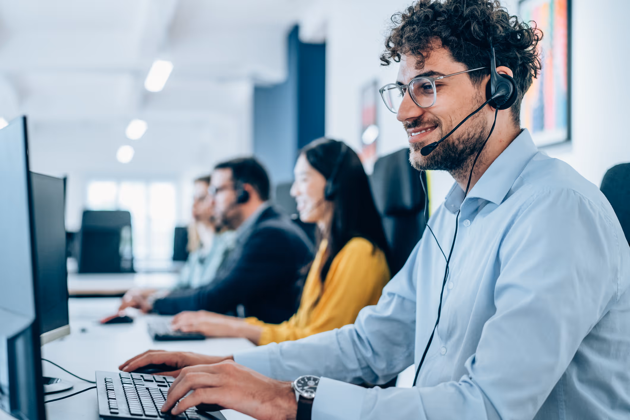 Man wearing headset and glasses typing on a keyboard in a modern call center with colleagues in the background.