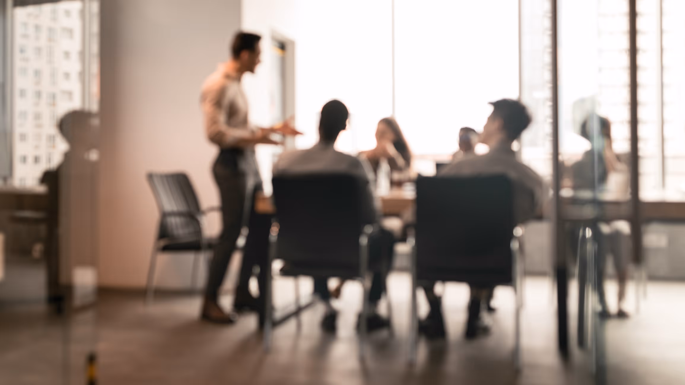 Blurred image of people seated around a conference table in an office with one person standing and speaking.