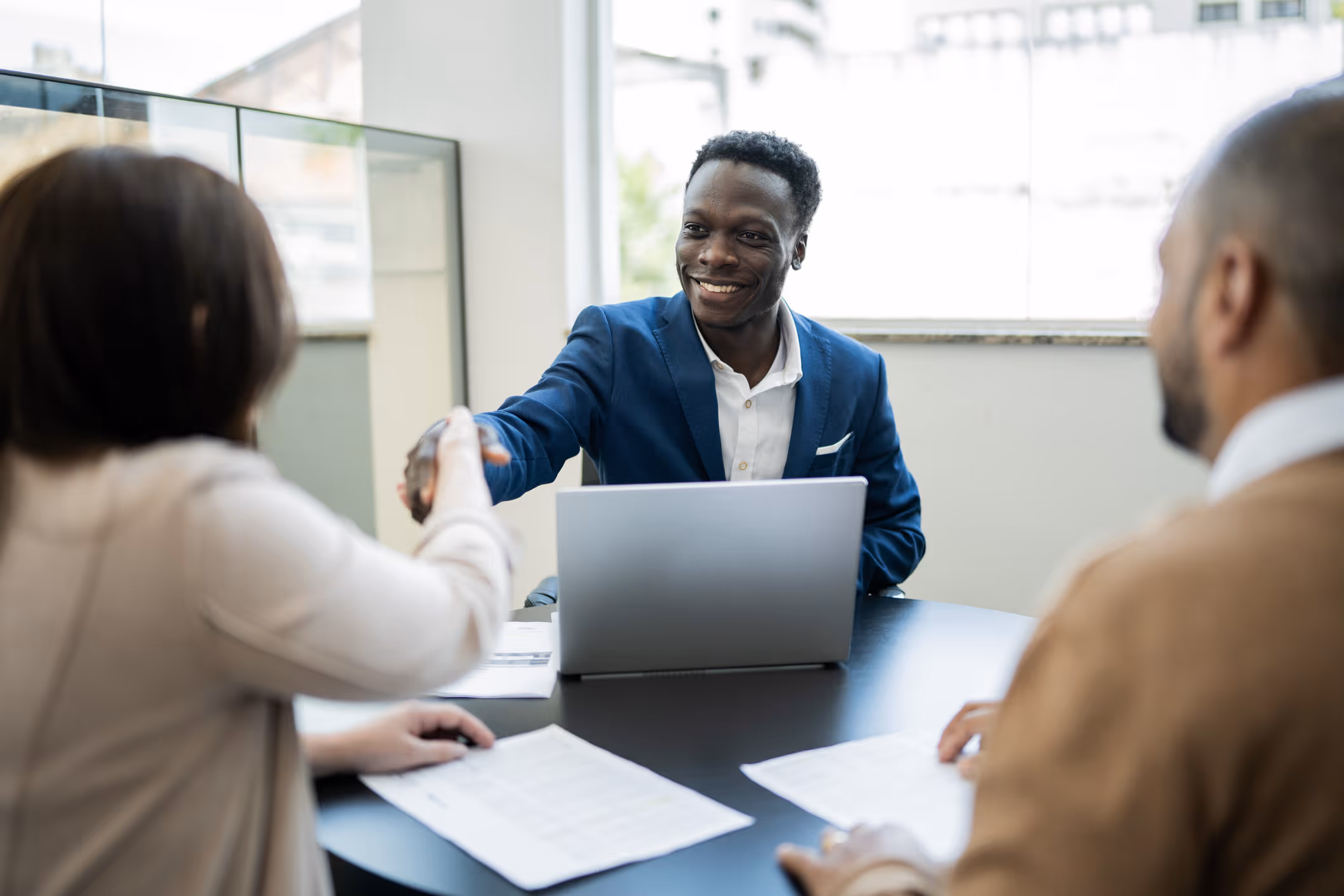 Smiling businessman shaking hands with a woman across a table with documents and a laptop.