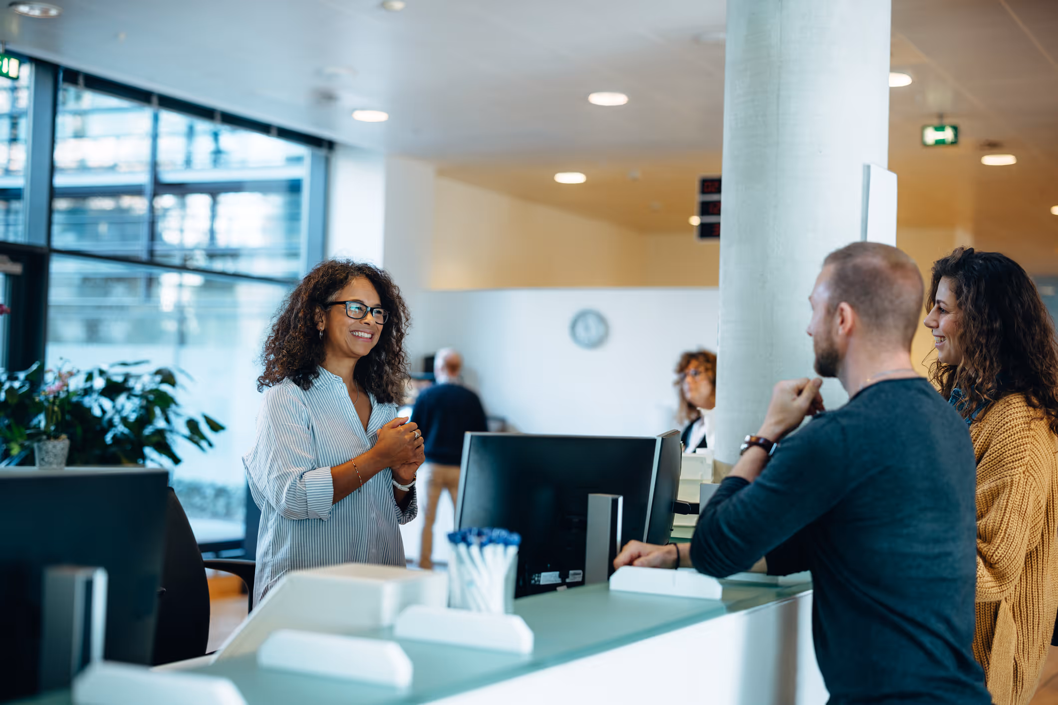 Receptionist smiling and assisting a man and woman at a modern office front desk.