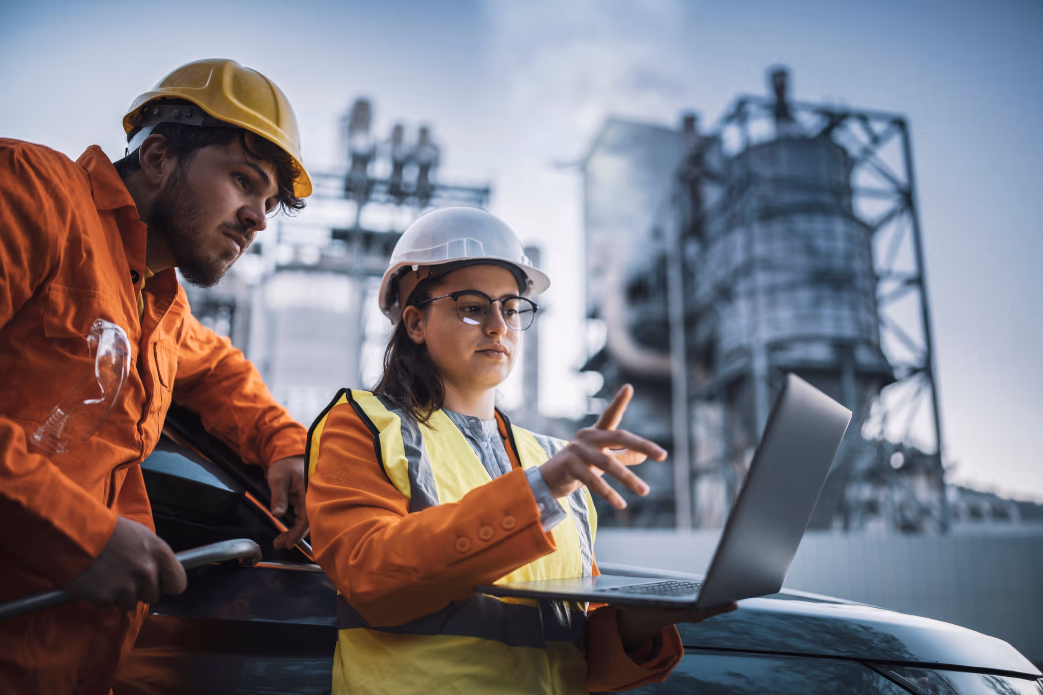 Two industrial workers wearing hard hats and safety gear review information on a laptop outdoors near a factory.