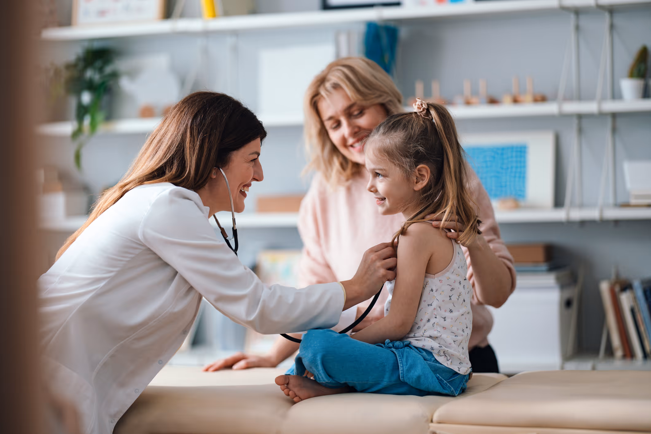 Female doctor using a stethoscope to examine a smiling young girl sitting on an exam table with her mother watching.