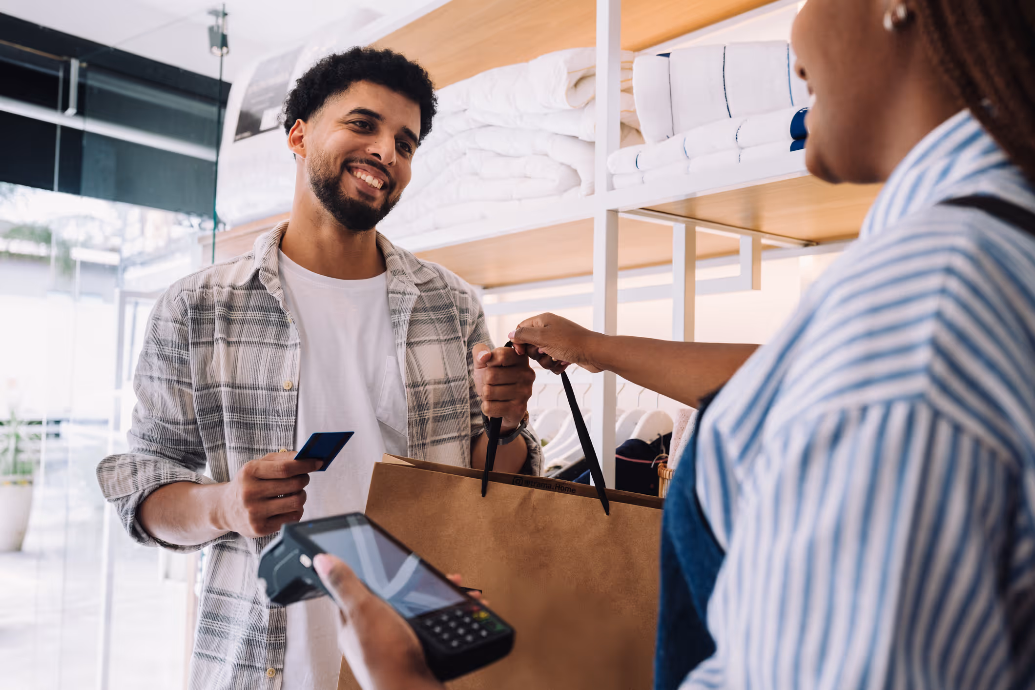 Smiling man holding a credit card and shopping bag while cashier processes payment using a card reader.
