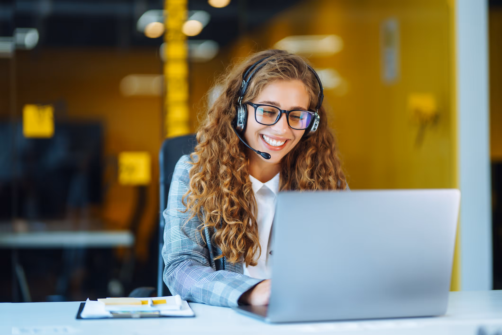 Smiling woman with curly hair wearing glasses and a headset, working on a laptop in an office.