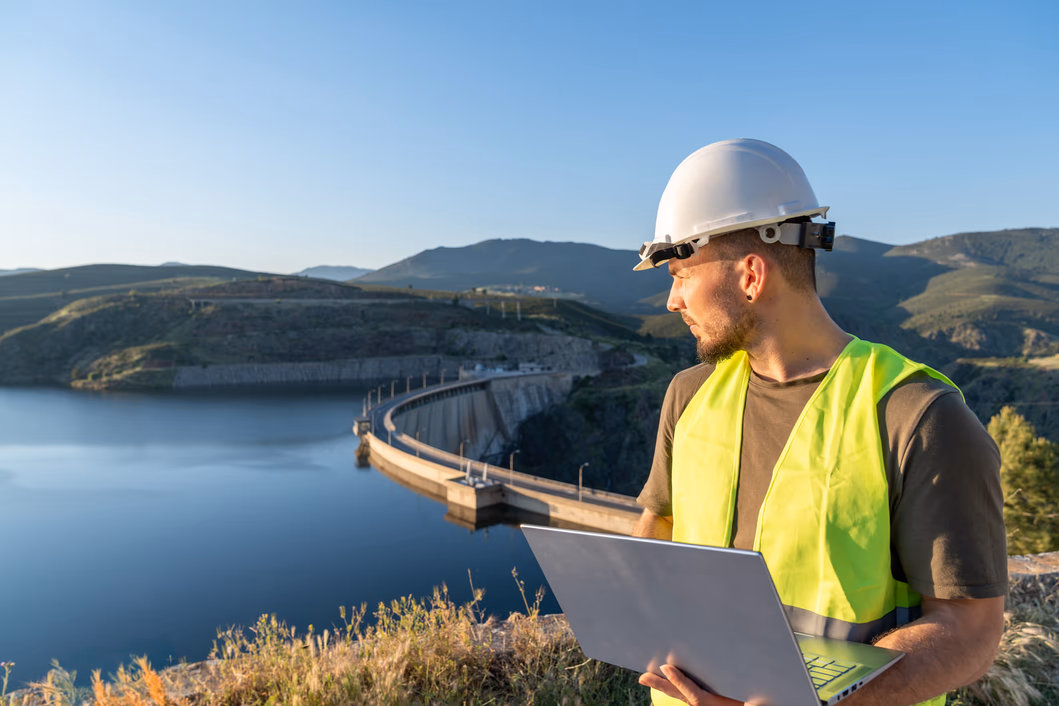 Engineer wearing a white hard hat and yellow safety vest holding a laptop, overlooking a dam and reservoir surrounded by hills.