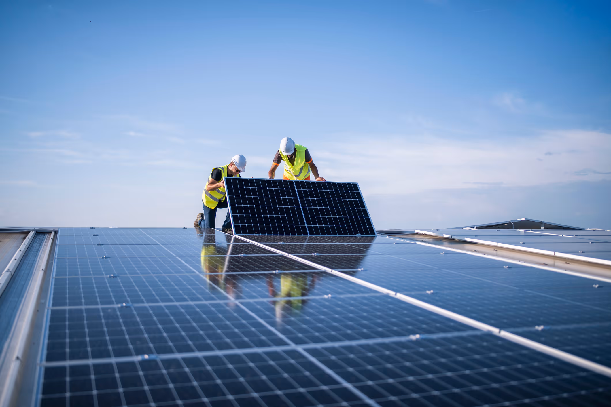 Two workers in safety vests and helmets installing solar panels on a roof under a clear blue sky.