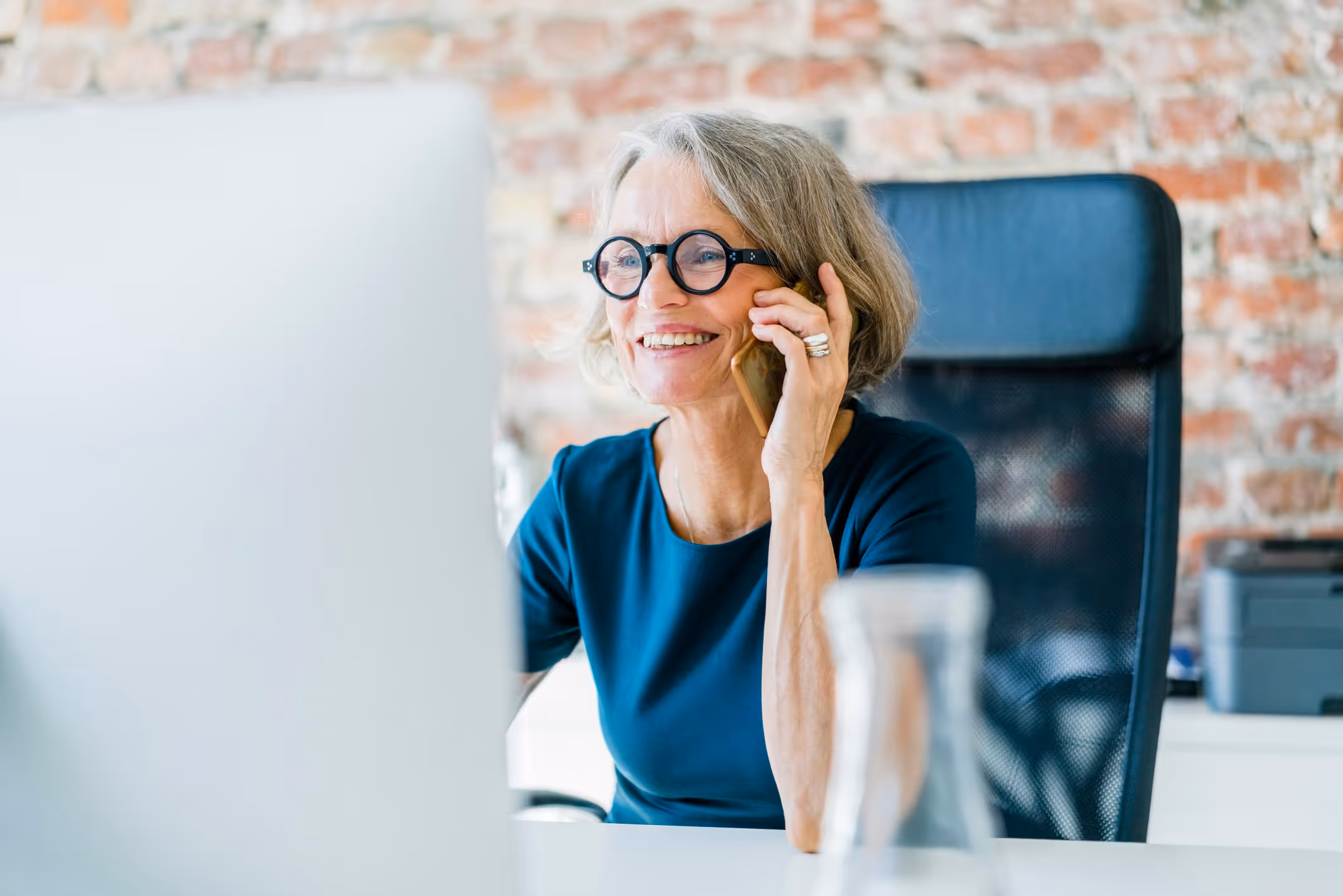 Smiling elderly woman with glasses talking on a smartphone while sitting at a desk in front of a computer.