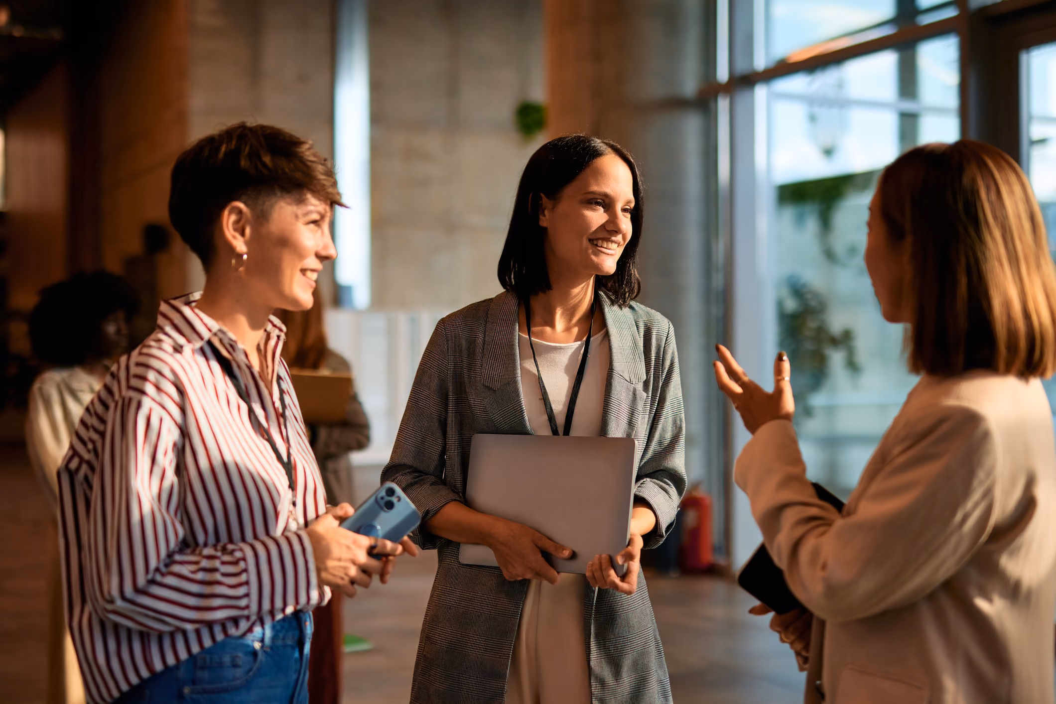 Three women smiling and chatting indoors, one holding a laptop and another holding a phone.