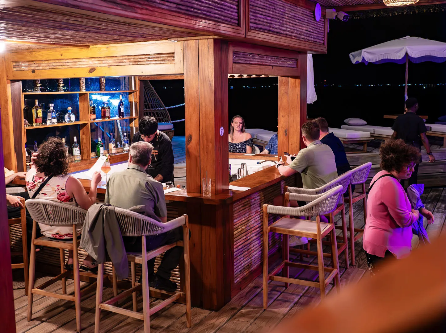 People sitting at an outdoor bar at night with a bartender serving drinks.