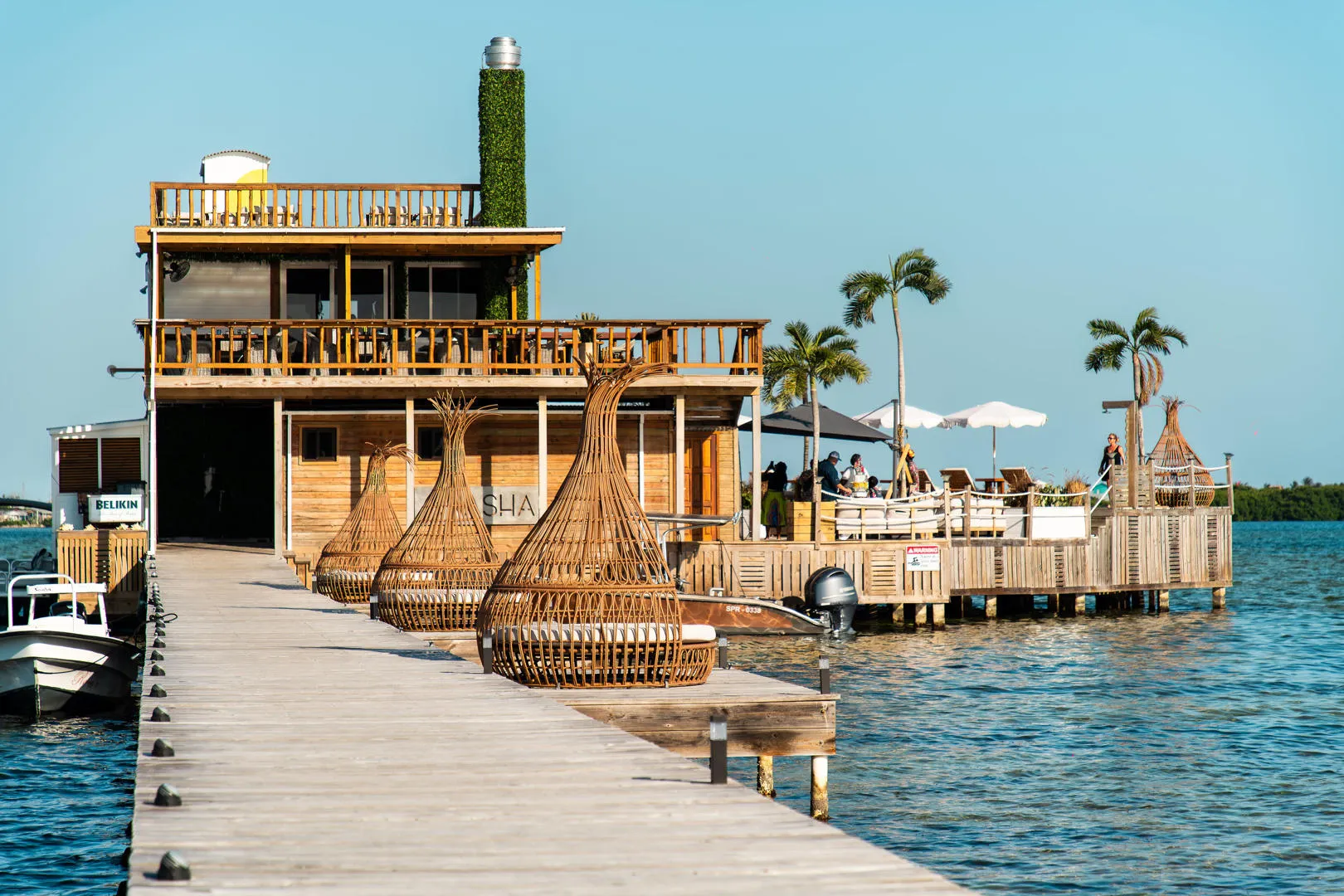 Wooden dock leading to waterfront restaurant with outdoor seating and palm trees.