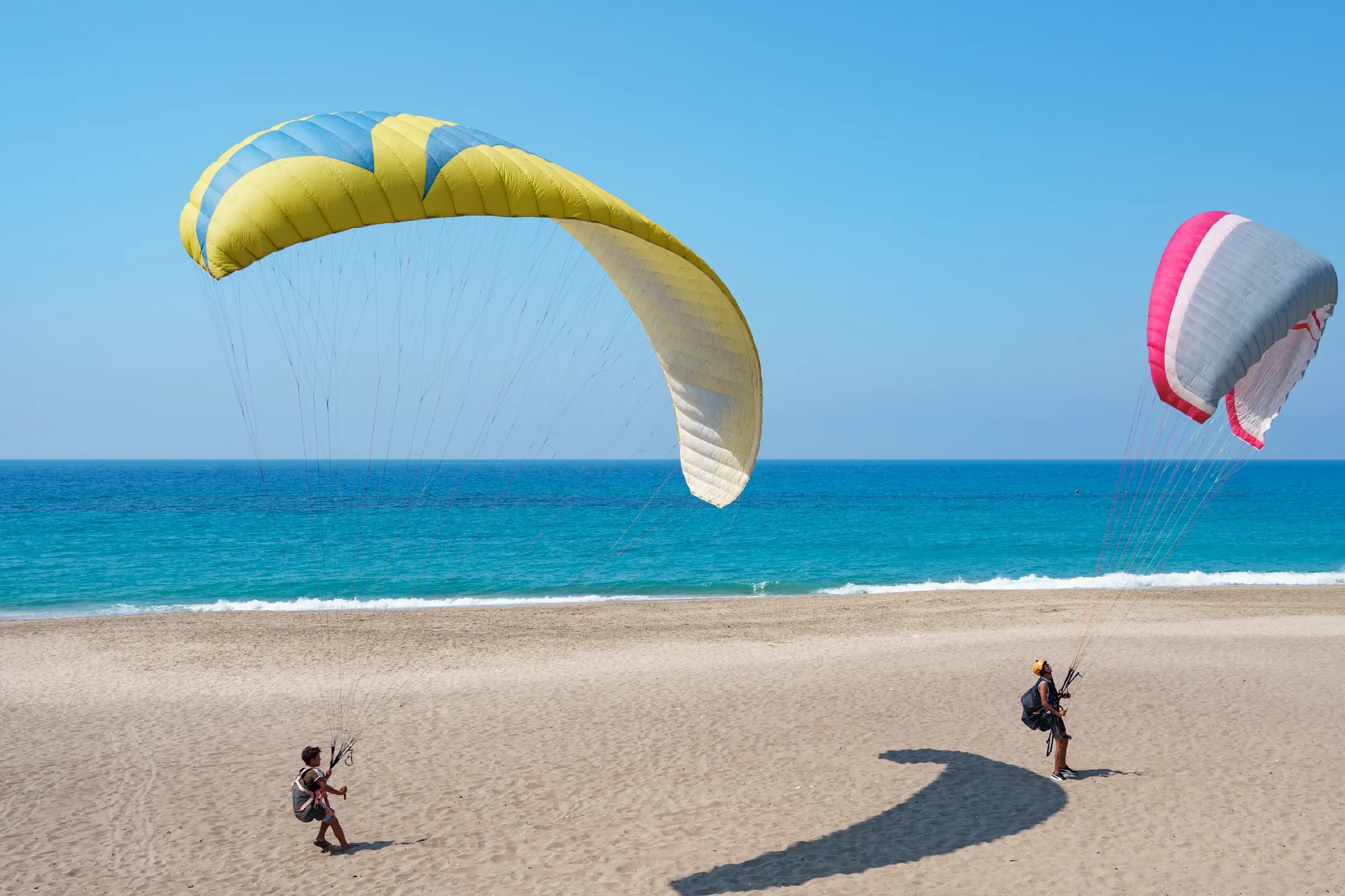 Two people paragliding on a sandy beach beside the ocean under clear blue skies