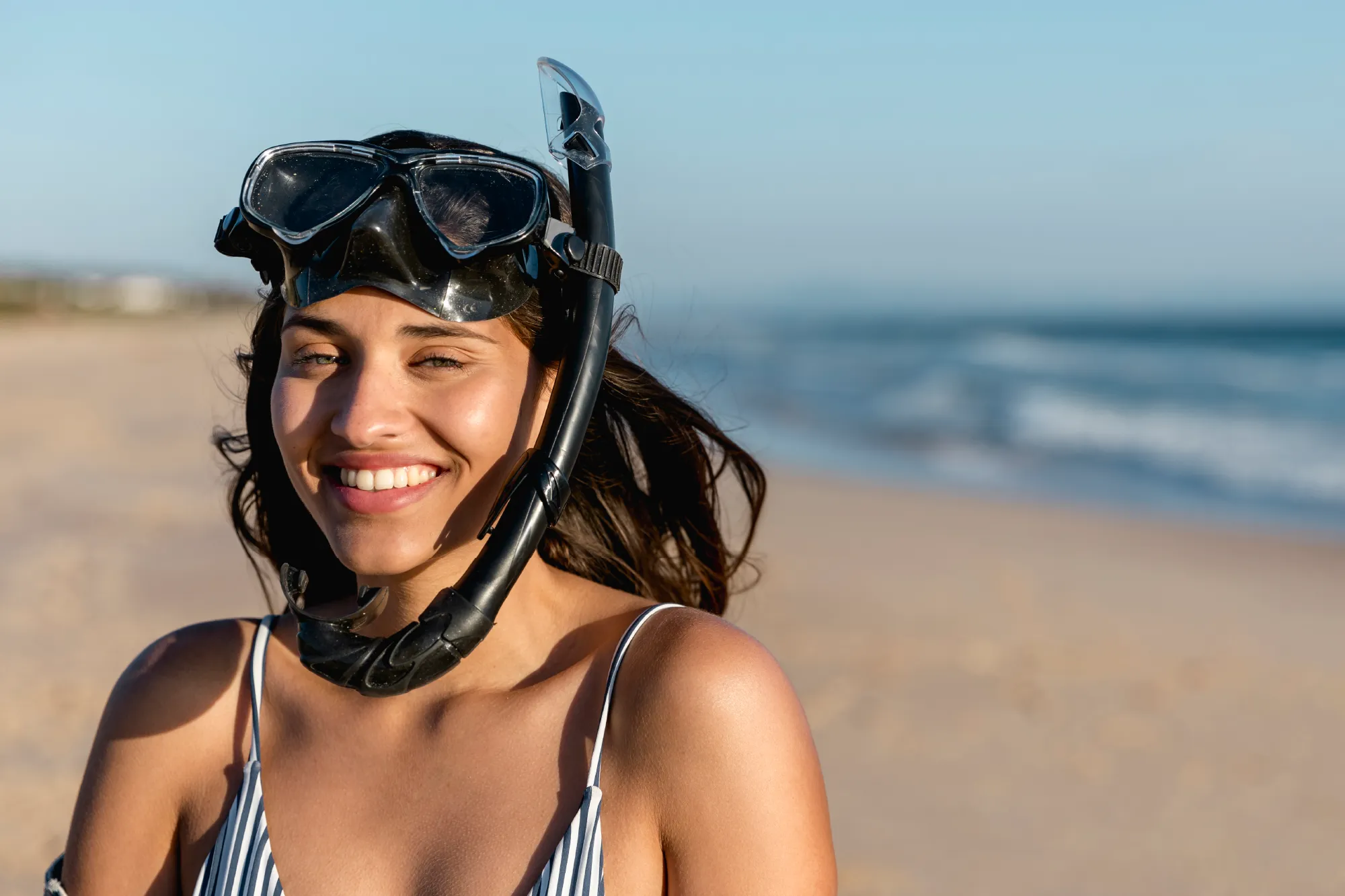 Smiling woman wearing a snorkel mask on a sunny beach with ocean waves in the background