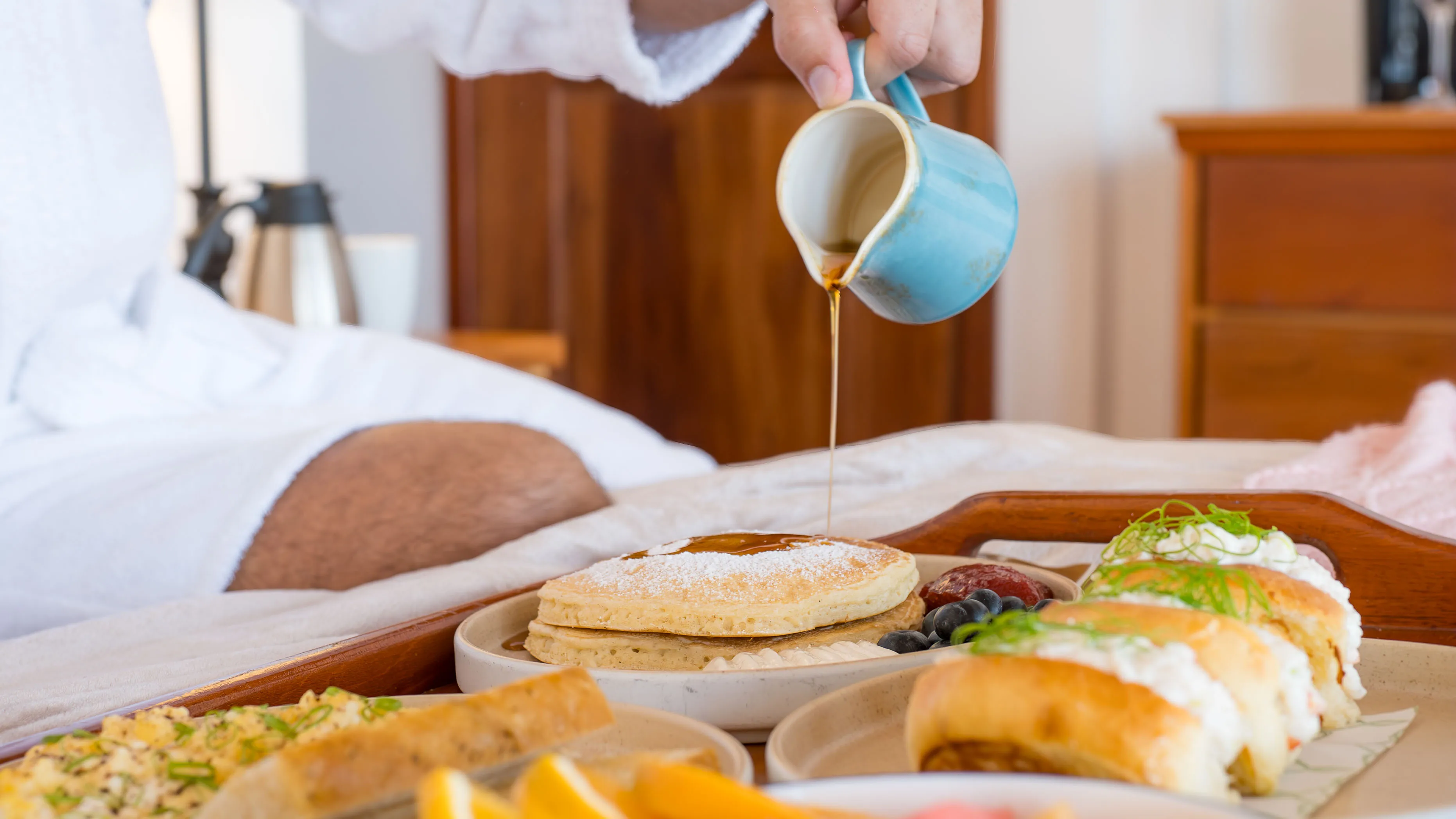 Breakfast in bed with pancakes, fruit, and coffee being poured in a cozy setting.