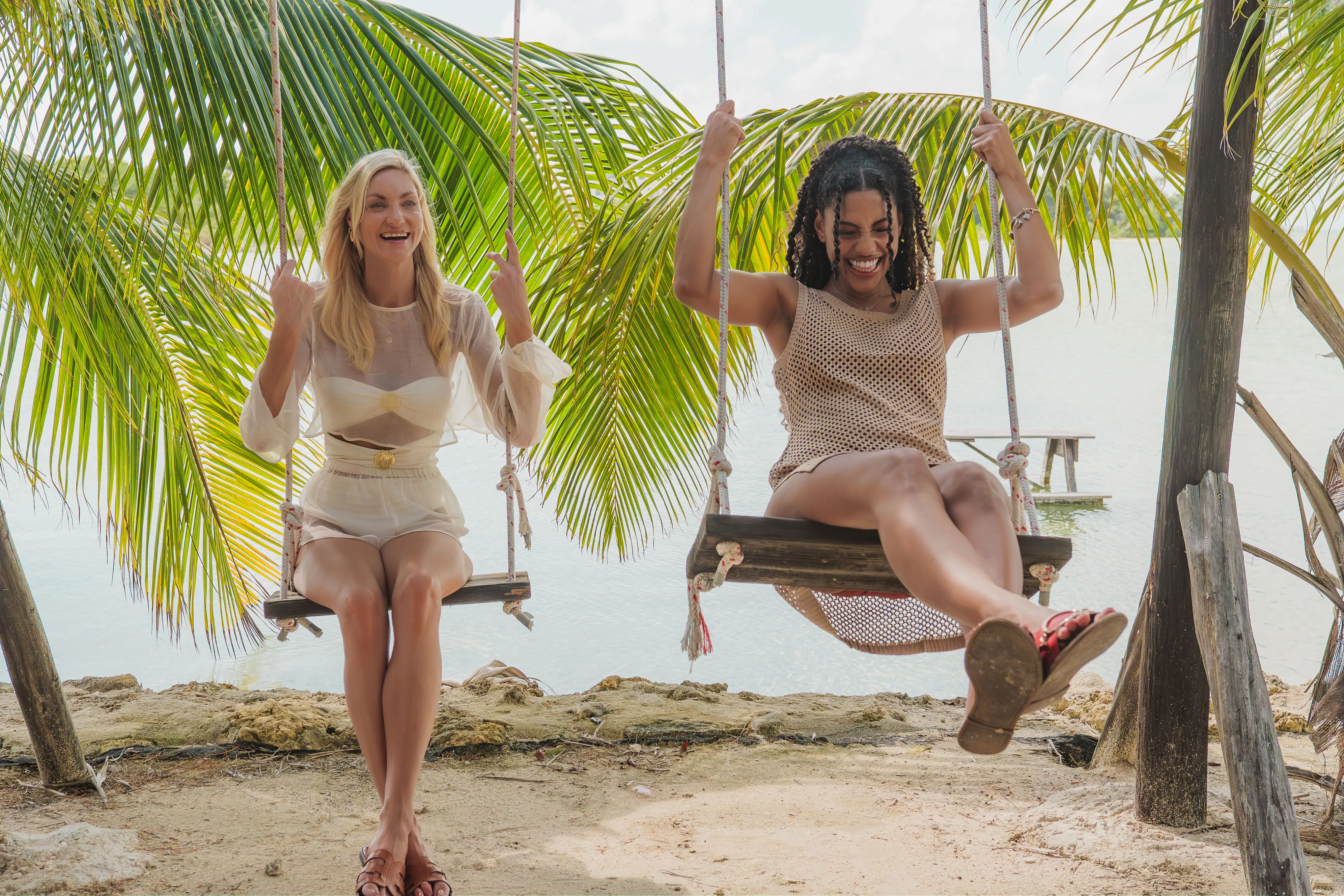 Two women enjoying a beach swing under palm trees with ocean views in the background.