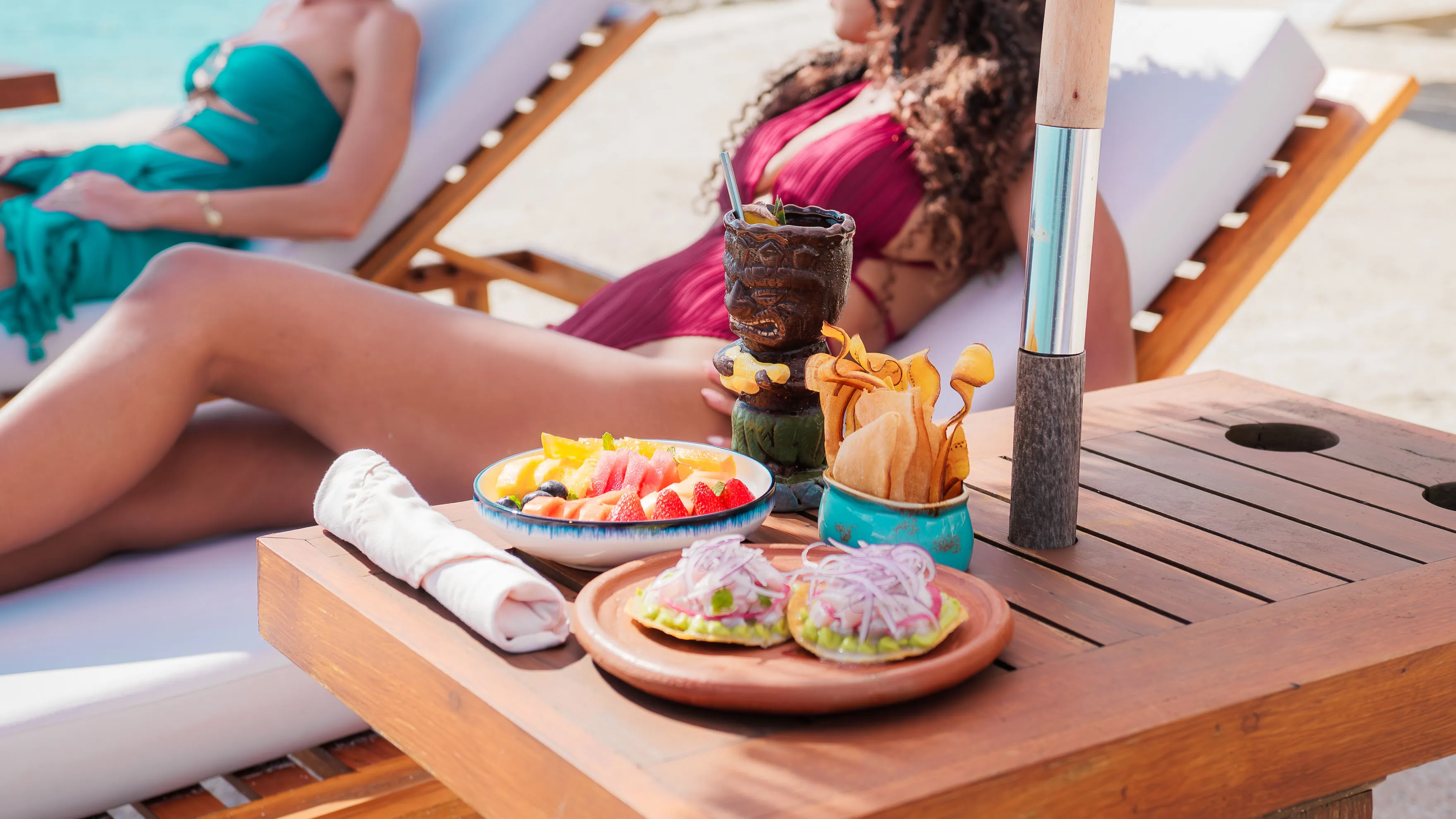 Poolside snacks with fruit bowl, chips, and tiki cocktail beside loungers