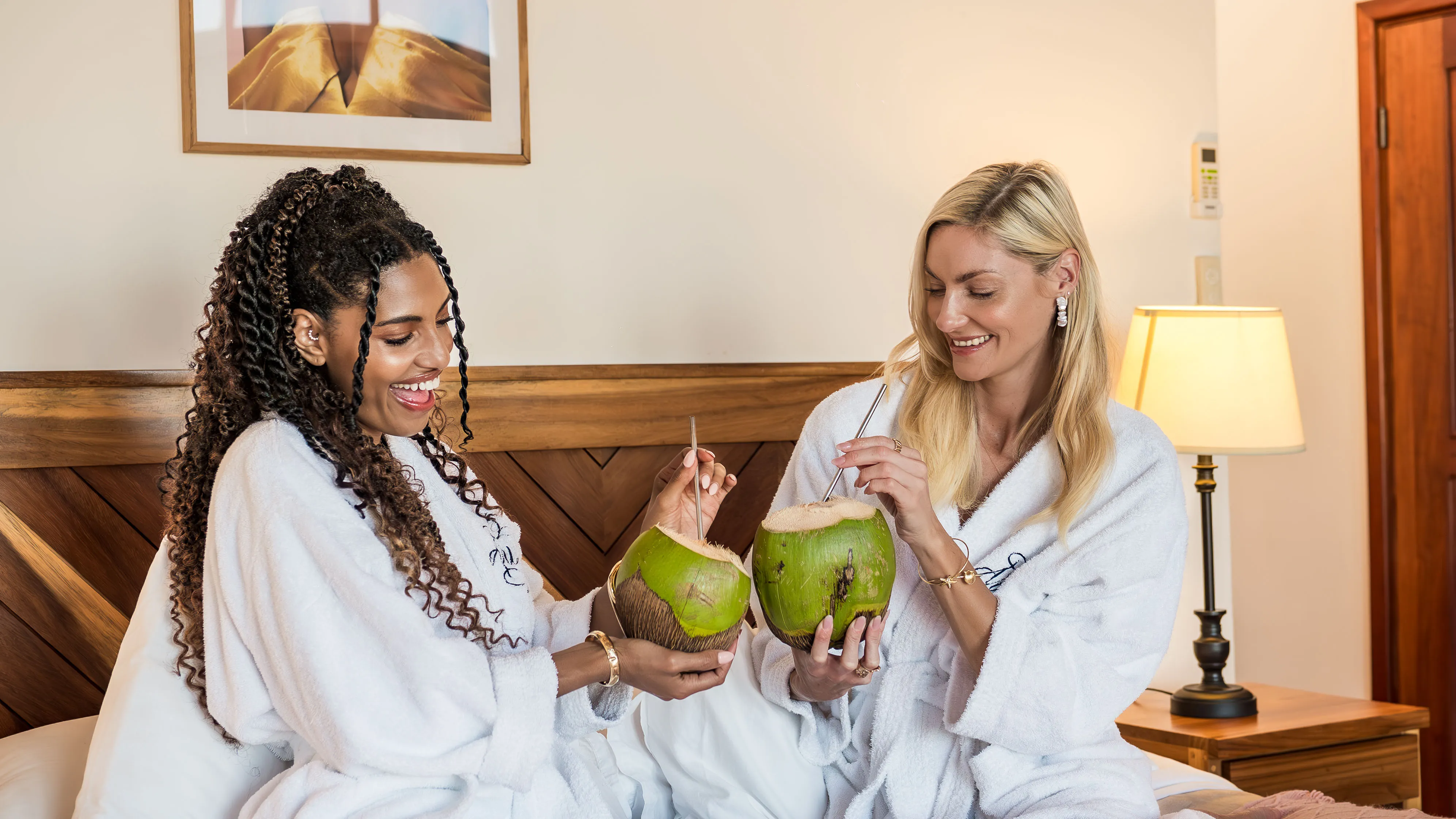 Two women in robes smiling and drinking fresh coconut water inside a villa.