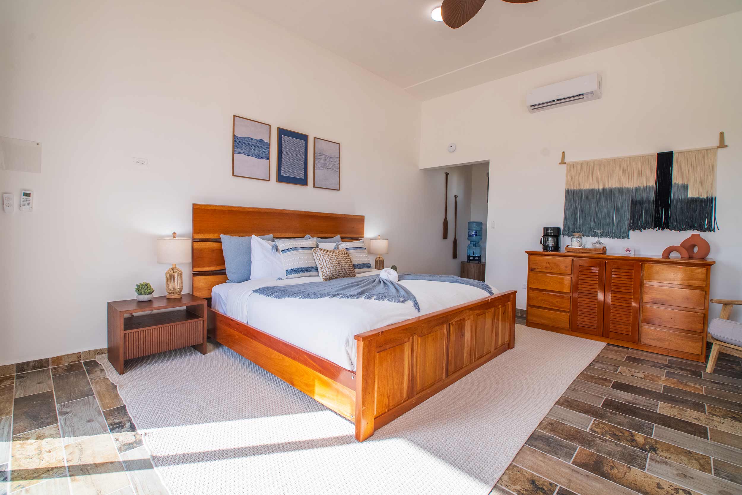 Spacious villa bedroom with a wooden king bed, neutral decor, and woven wall art in a bright, airy interior.