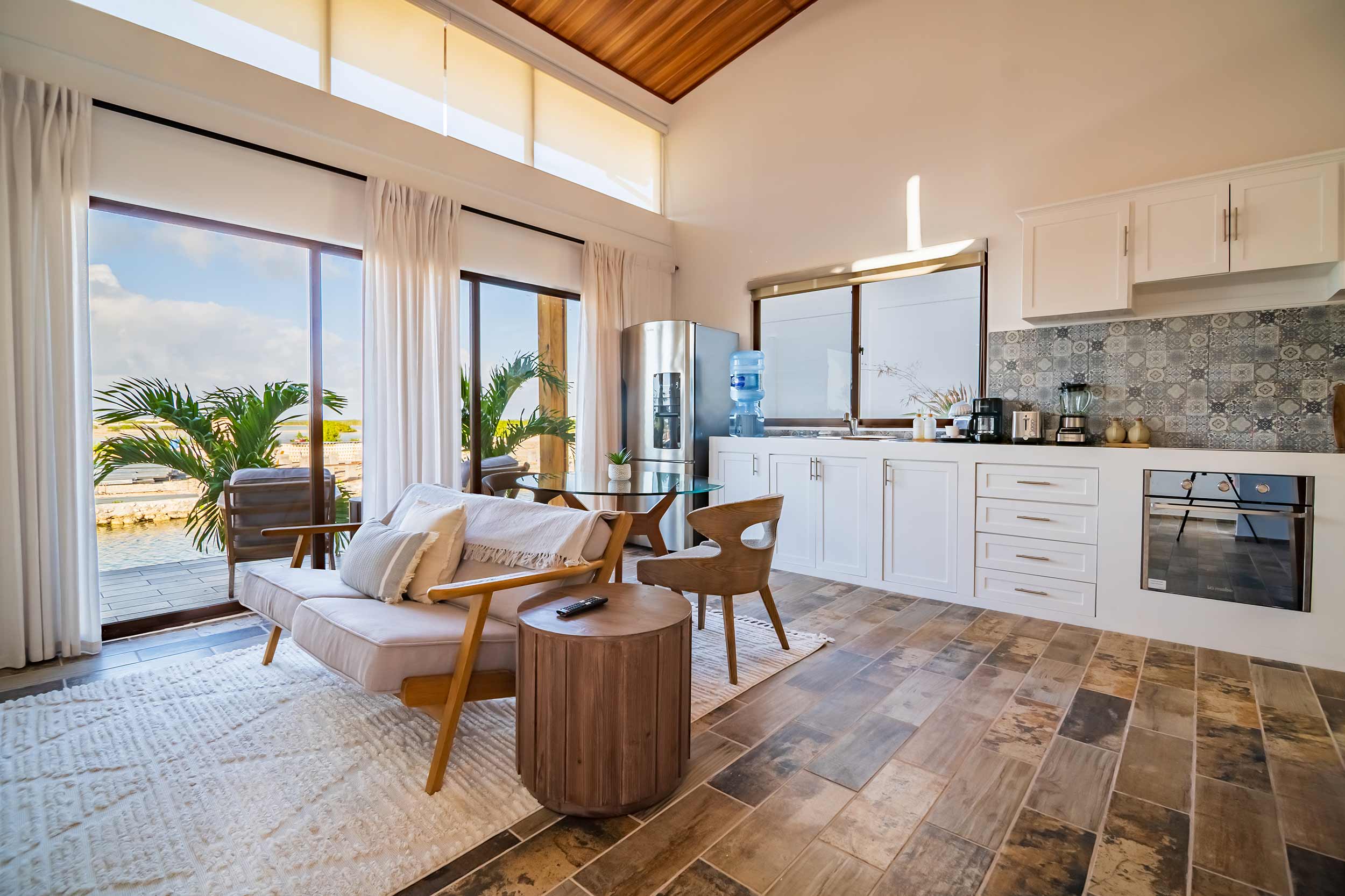 Open-concept villa interior with living room seating, full kitchen, and large windows overlooking tropical scenery.
