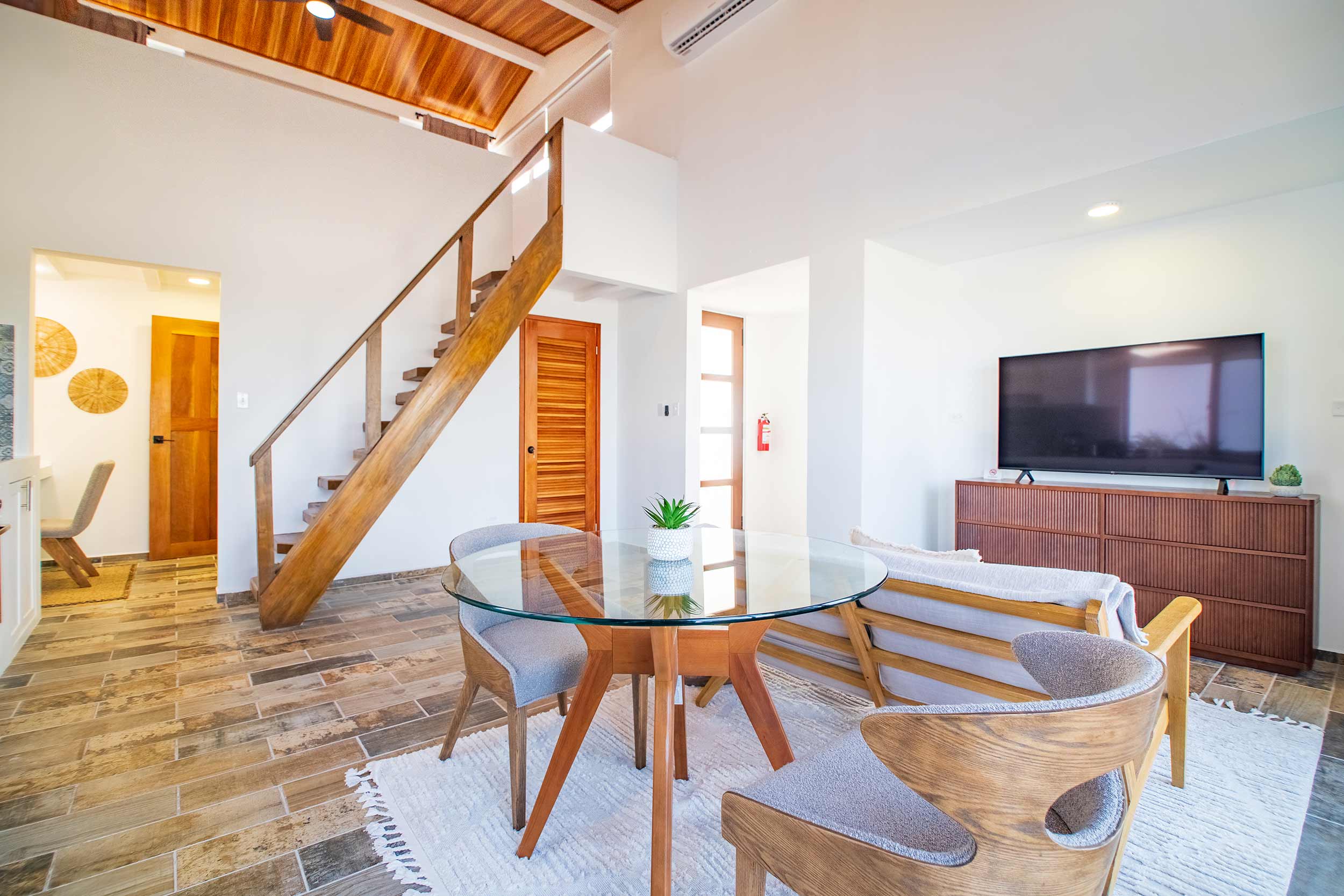 Bright villa living area with round glass dining table, wooden staircase, and TV lounge space.
