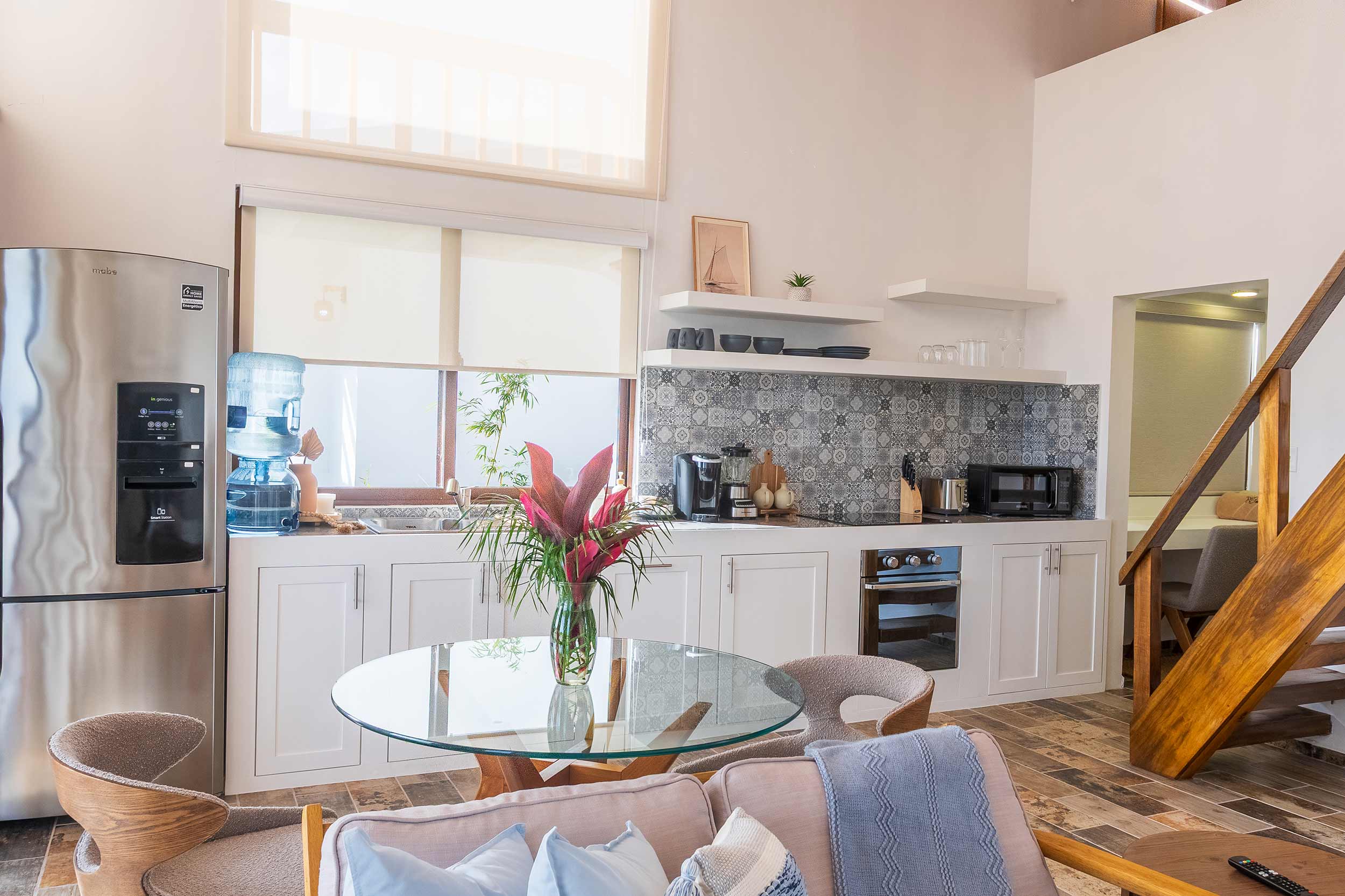 Bright open-concept villa kitchen and dining area with patterned tile backsplash, glass dining table, and wooden staircase.