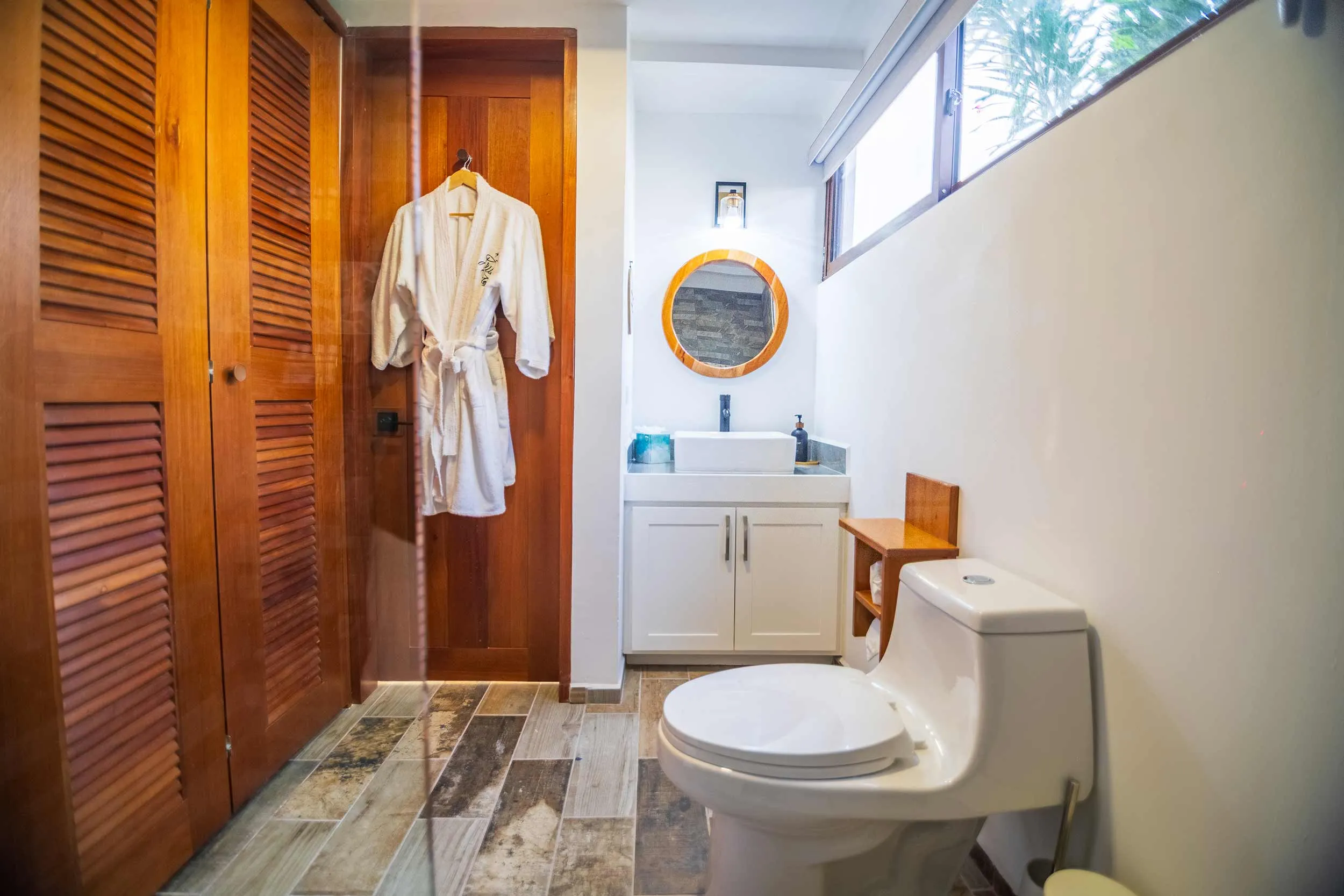 Villa bathroom with white vanity sink, round mirror, wooden doors, and hanging bathrobe.
