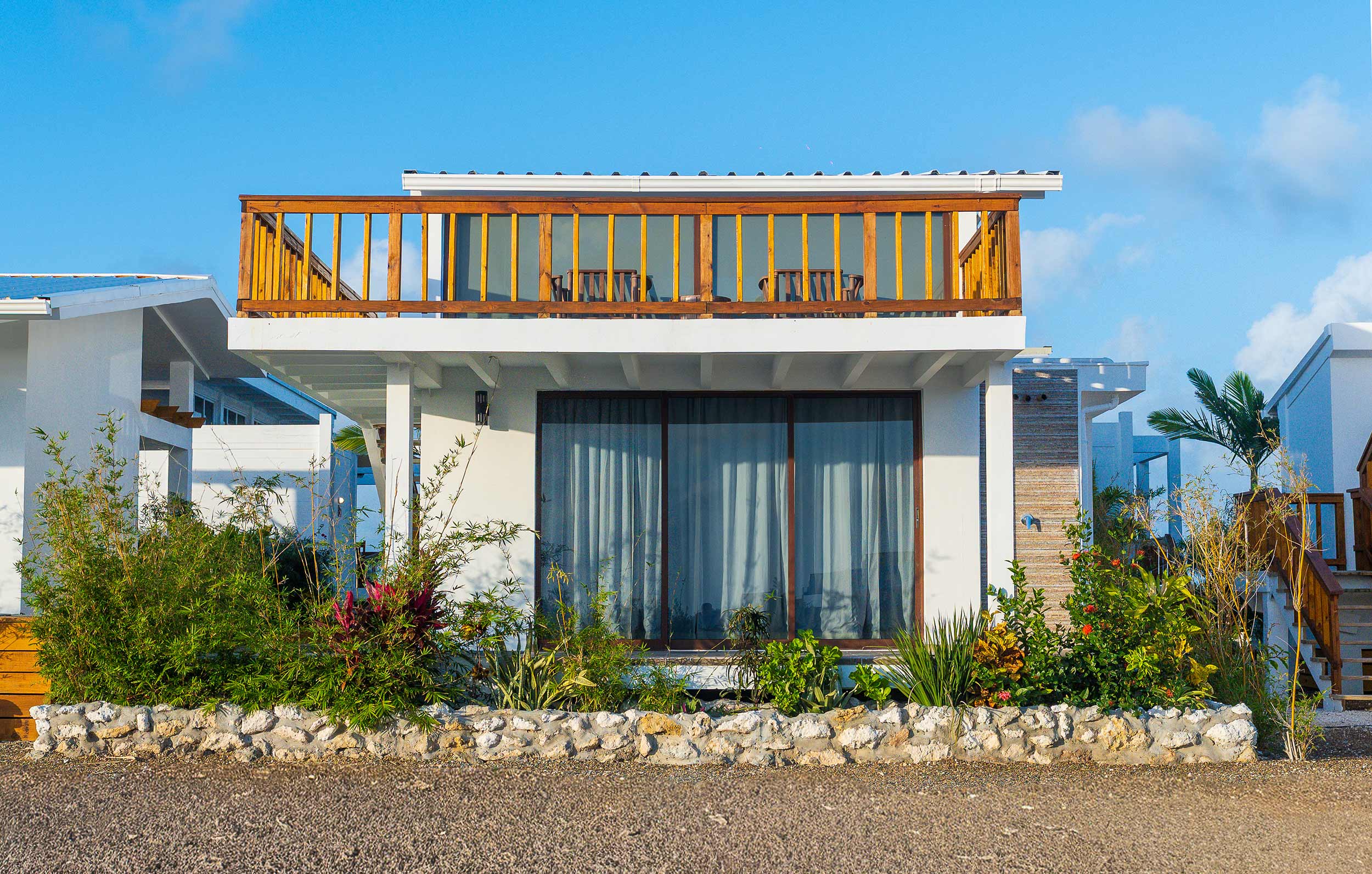 Modern beachfront villa exterior with rooftop balcony, tropical landscaping, and large glass doors.
