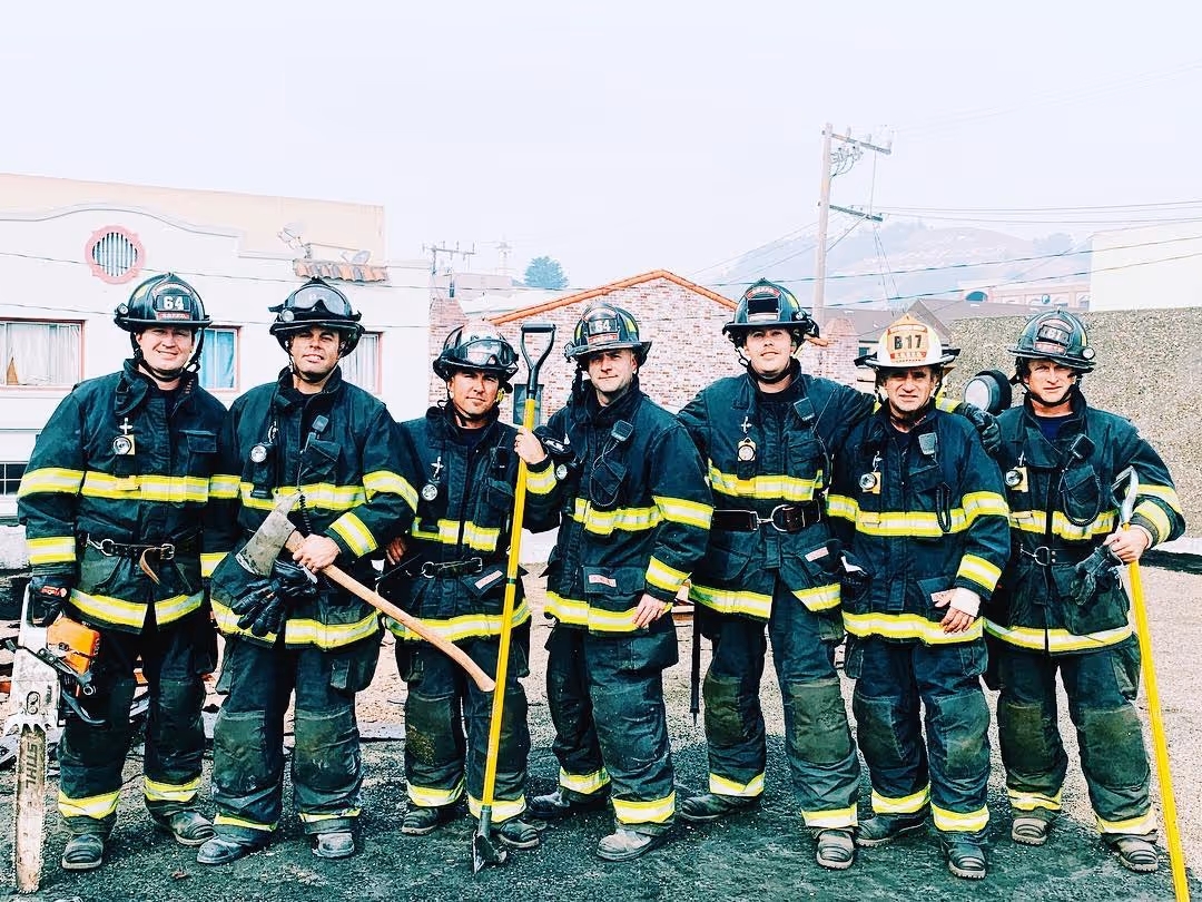 Seven firefighters in full gear posing together outdoors with firefighting tools.