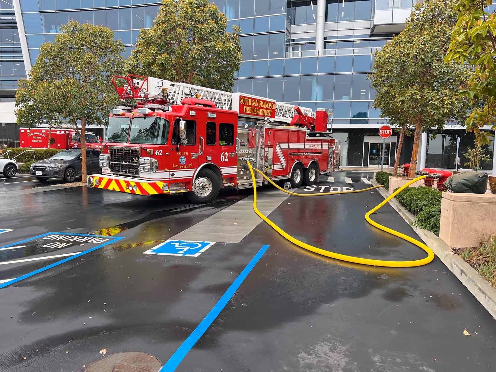 Red South San Francisco fire truck with ladder parked near building, connected to a yellow fire hose on wet pavement.