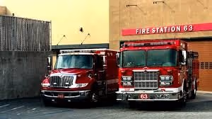 Two red fire trucks parked outside Fire Station 63 building with a beige wall.