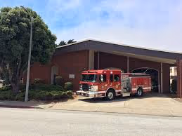 Red fire truck parked outside a fire station with brick walls and a large tree nearby.
