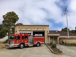 Red fire truck parked in front of a beige fire station building with a flagpole and trees in the background.