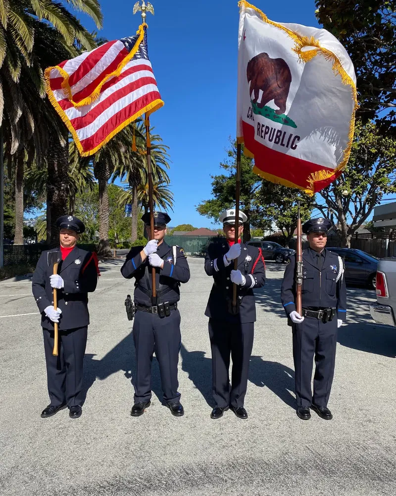 Four uniformed officers standing in a row holding American and California Republic flags and an axe on a sunny street with palm trees.