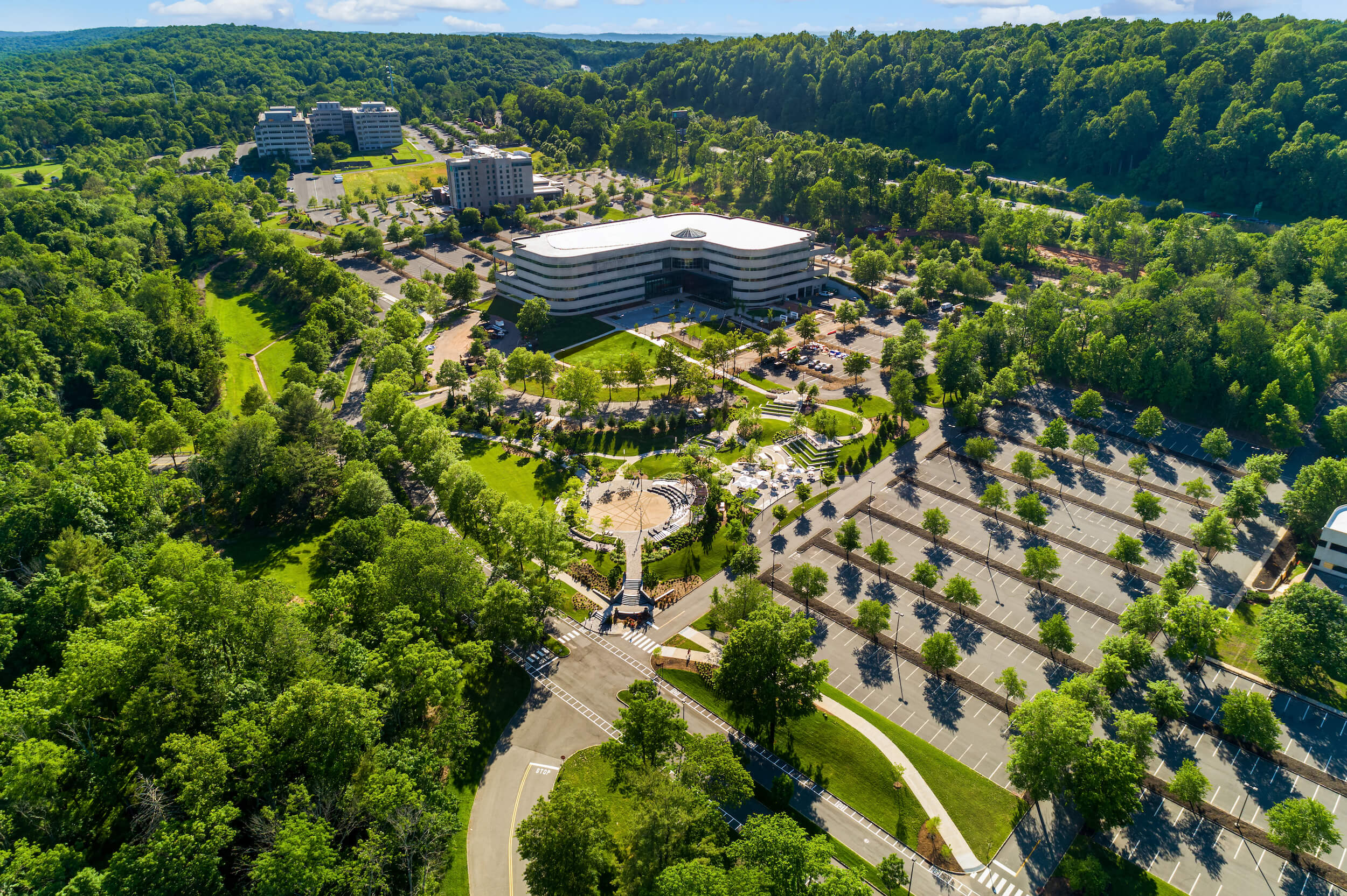 Aerial view of a modern office building surrounded by green trees, landscaped gardens, and large parking lots under a clear sky.