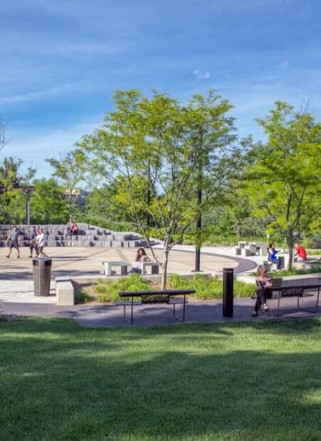 Park scene with green grass, trees, benches, and people sitting or walking on a paved area under a blue sky.