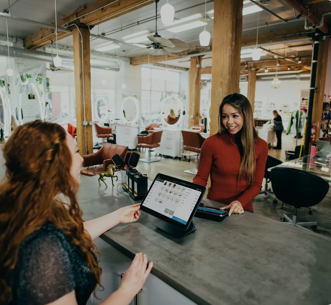 A smiling woman in a red sweater assists a customer who is interacting with a touchscreen at a salon reception desk.