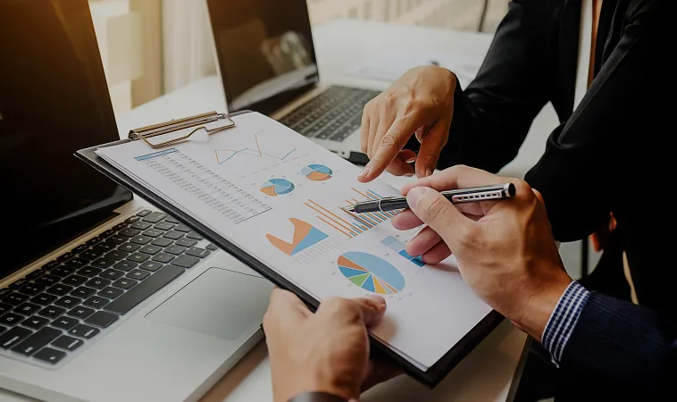 Two people reviewing financial charts and graphs on a clipboard next to a laptop.