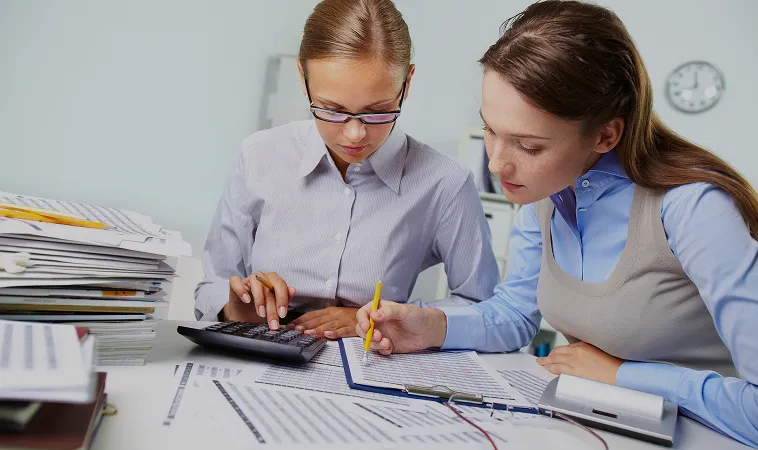 Two women working together on financial documents with a calculator and papers spread on a desk.
