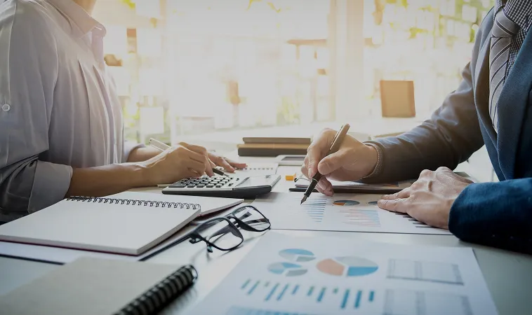 Two people working at a desk with financial charts, a calculator, notebooks, and eyeglasses in a bright office setting.