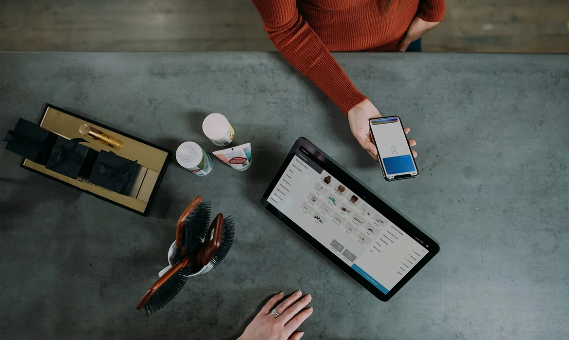 Overhead view of a person holding a smartphone with a payment screen next to a tablet displaying an online store, on a grey table with hairbrushes and cosmetic products.