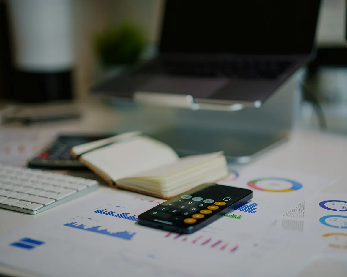 Workspace with smartphone calculator app open on screen, notebook, keyboard, laptop, and printed charts with graphs.