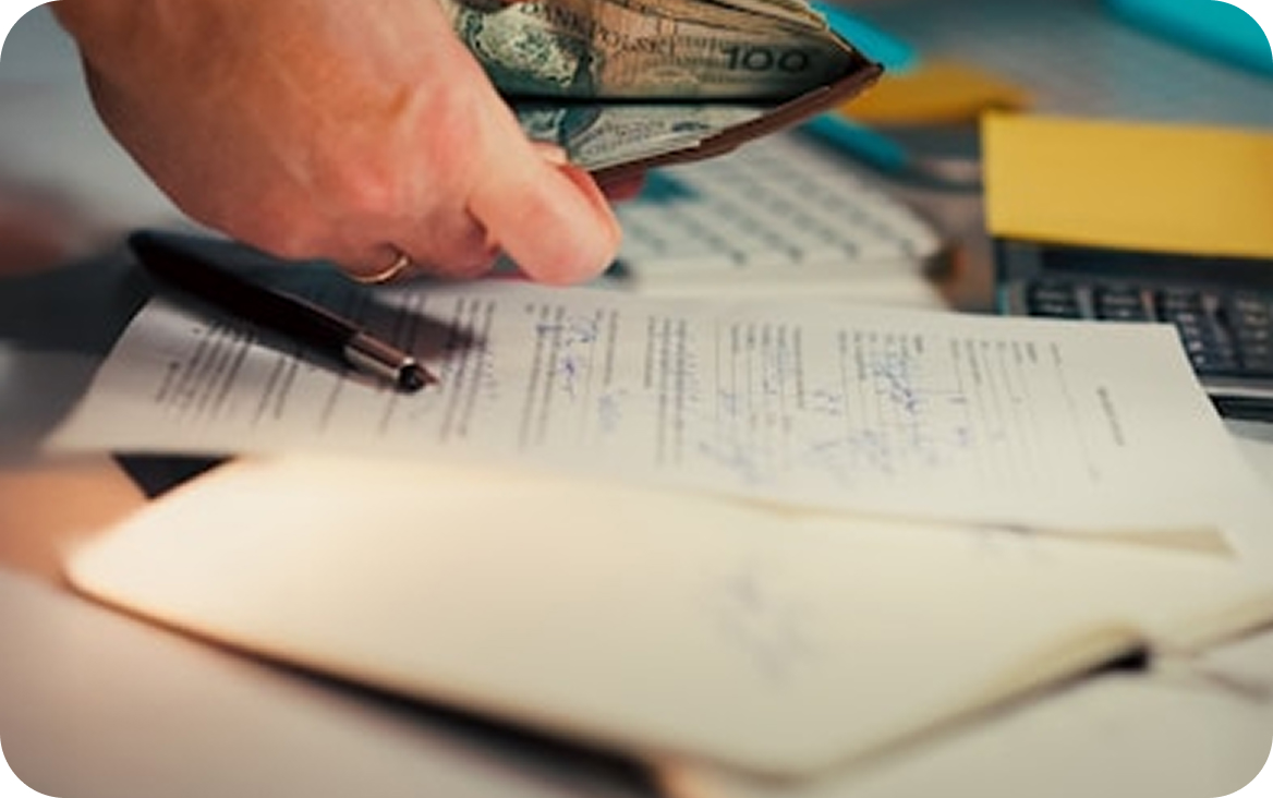 Hand holding an open wallet with cash above a signed contract, pen, and calculator on a desk.