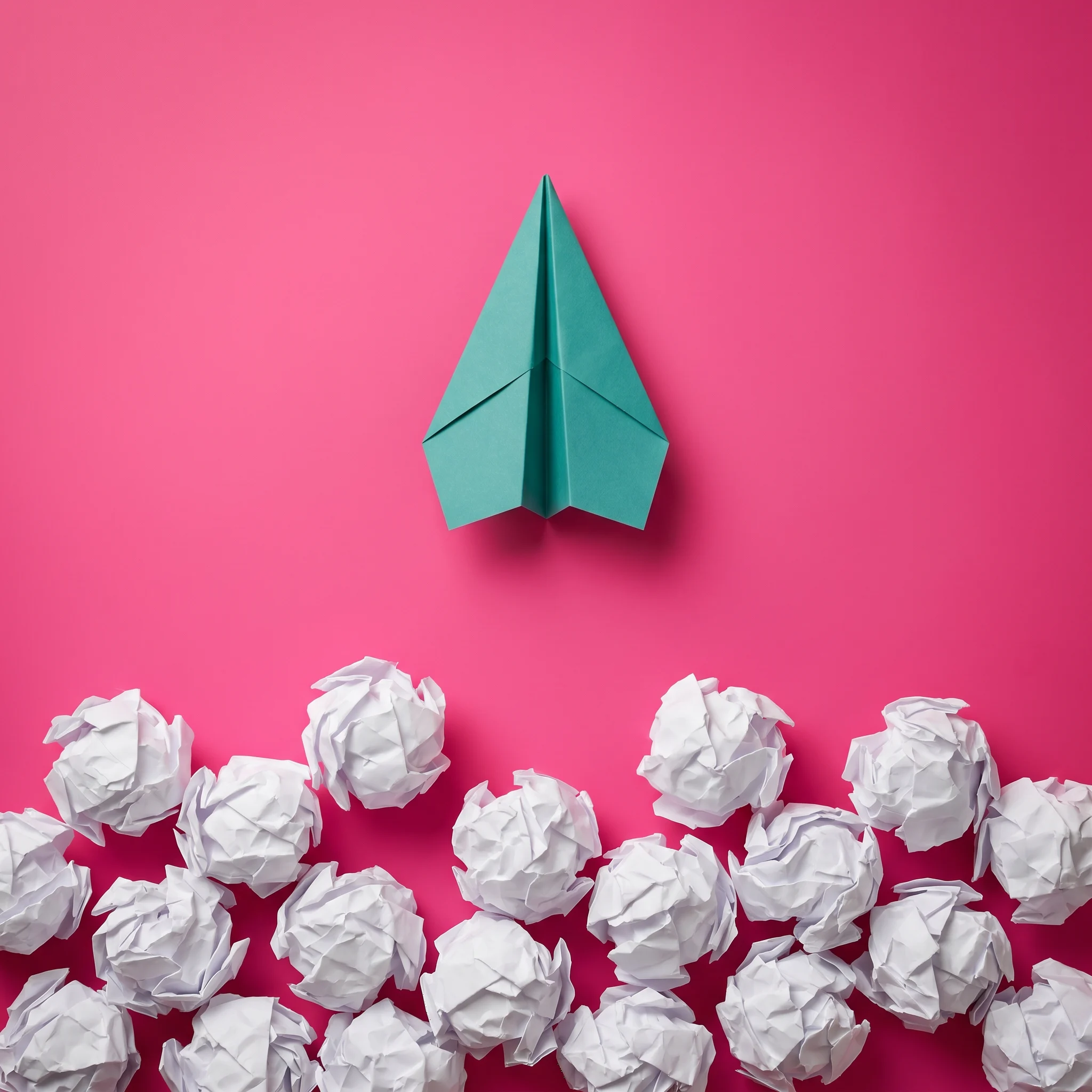 A green paper airplane above a group of crumpled white paper balls on a pink background, symbolizing creativity and standing out.