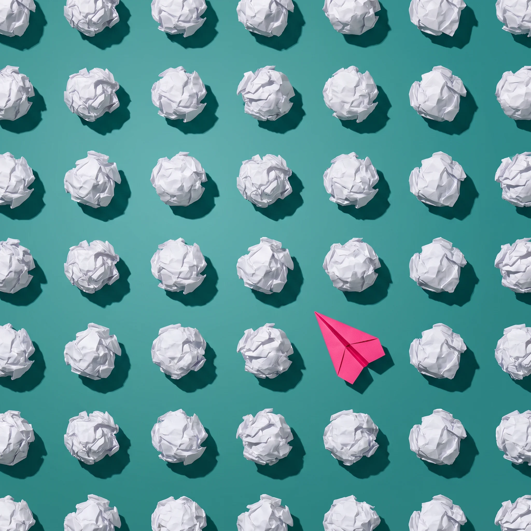 Grid pattern of white crumpled paper balls on a teal background with a single pink paper airplane among them.