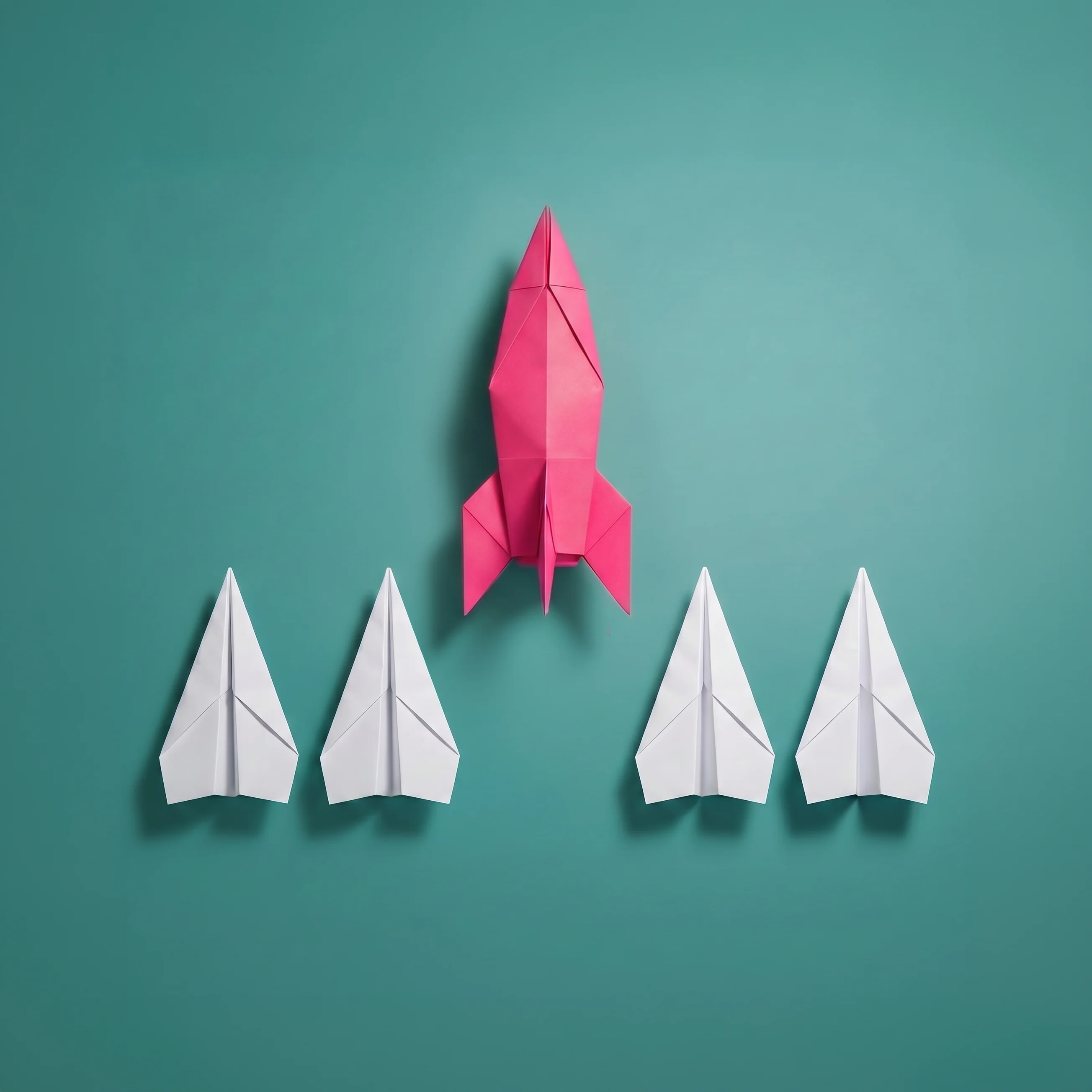 One pink paper rocket pointing upward surrounded by four white paper airplanes on a teal background, symbolizing leadership and standing out.