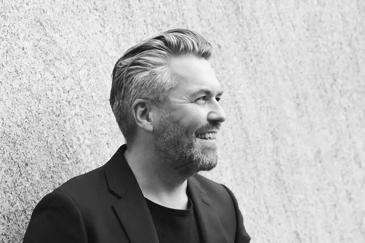 Black and white portrait of a smiling man with grey hair and beard, wearing a black blazer and shirt, looking to the right against a textured wall background.